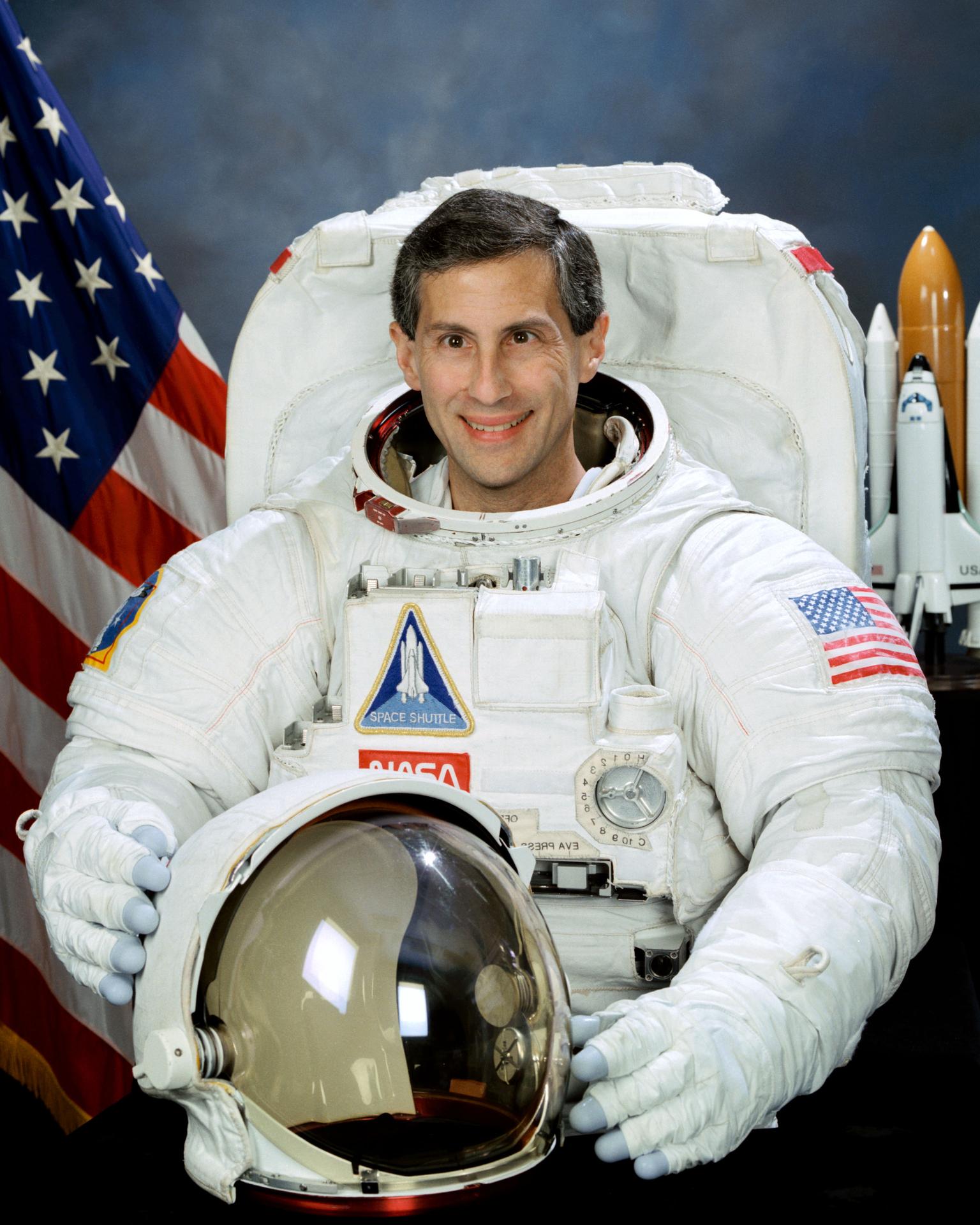 A man sits before the American flag in an astronaut suite, with his helmet off