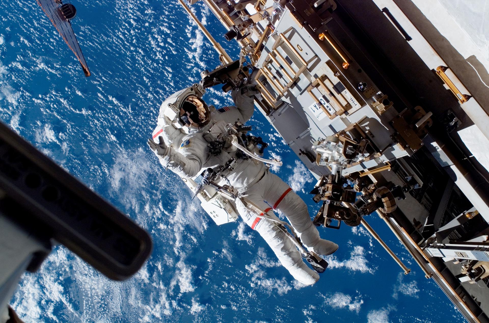 astronaut in space suit attached to the ISS floats with the earth far below them, working on the ISS 