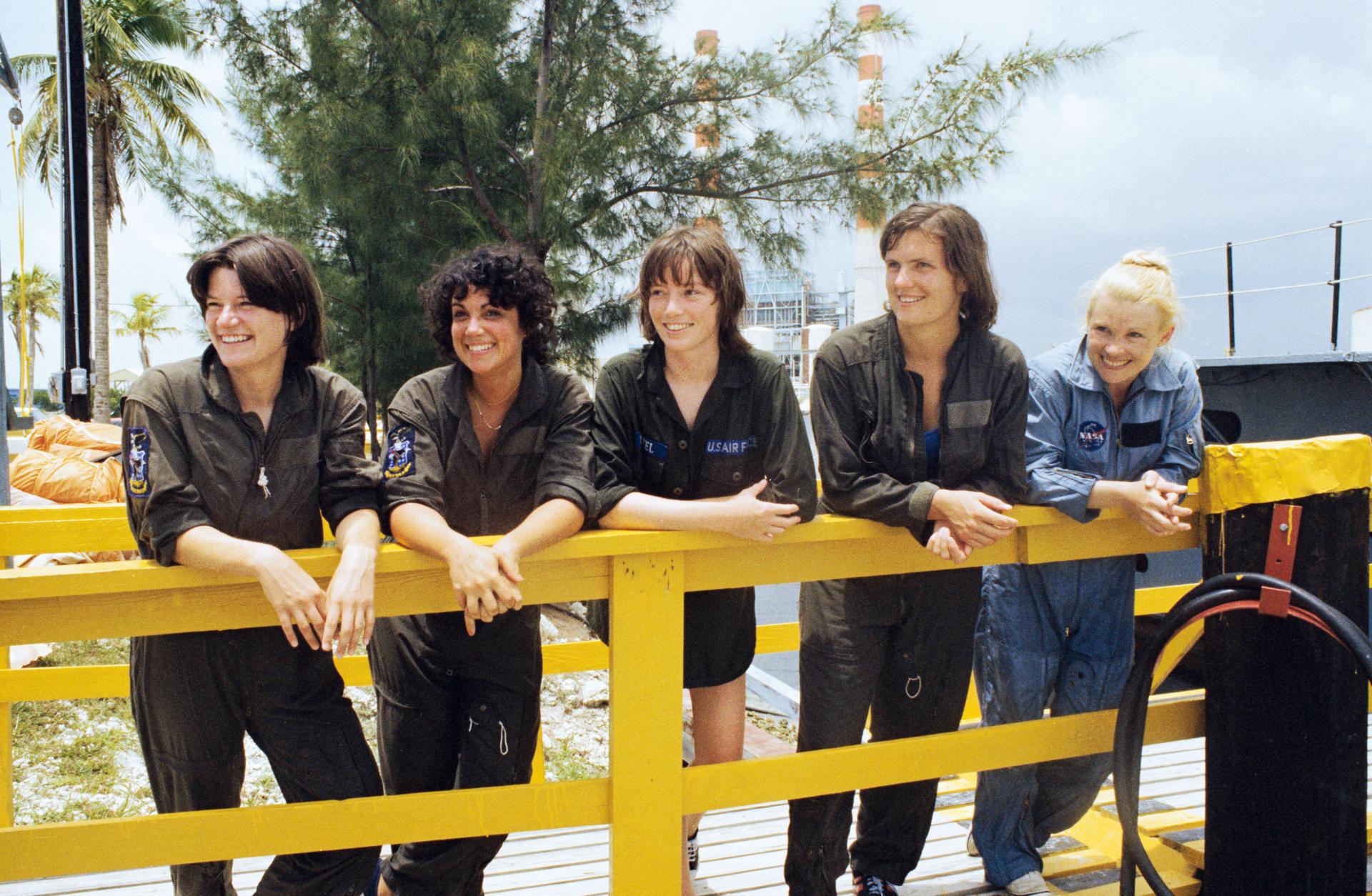 The first five women astronauts leaning up against a fence in green and blueoveralls.