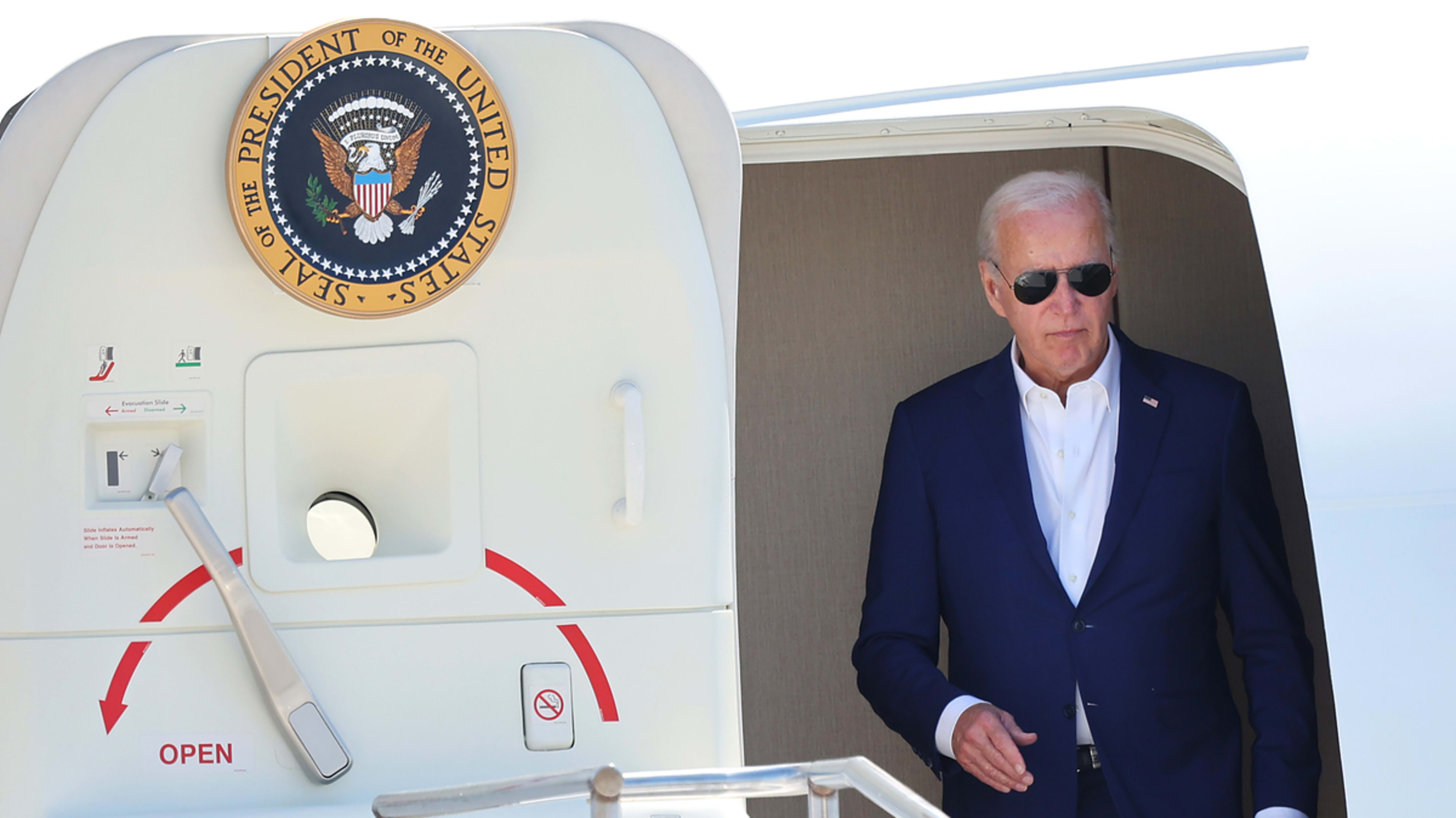 Joe Biden, wearing a dark suit and sunglasses, exits an aircraft