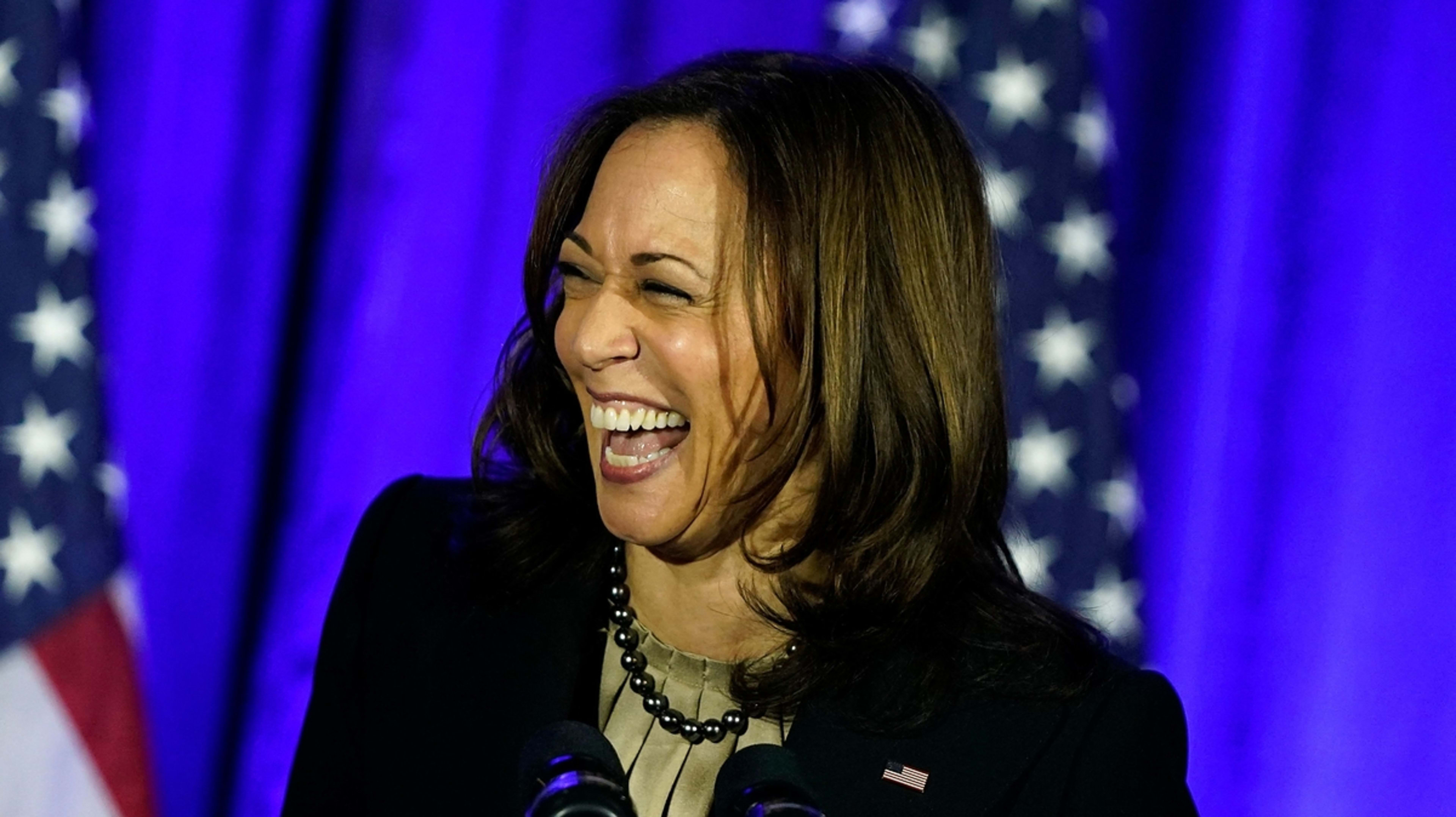 Vice President Kamala Harris laughing while speaking at an event, with two American flags in the background. She is wearing a dark blazer and a light top