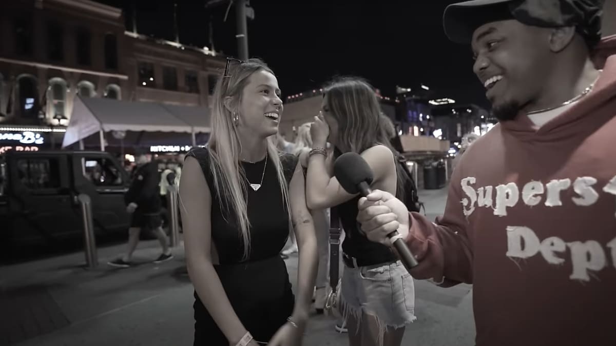 Two women are being interviewed outdoors at night by a man in a hoodie that reads "Superstar Dept" holding a microphone. They are smiling and laughing