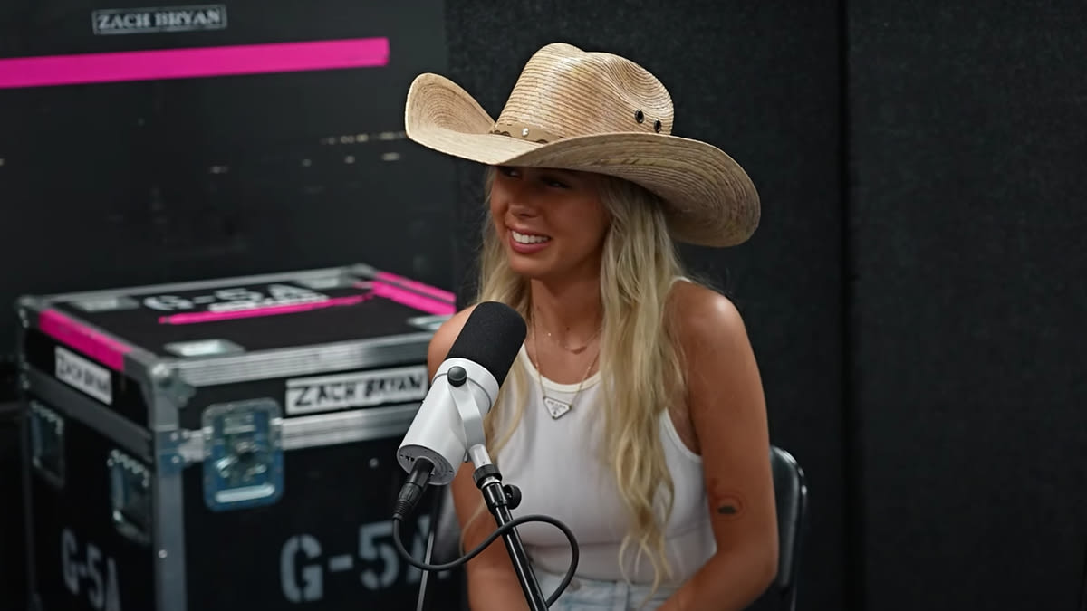 Person with long hair wearing a cowboy hat and sleeveless top sits at a microphone in a recording studio. Name on equipment reads "Zach Bryan."