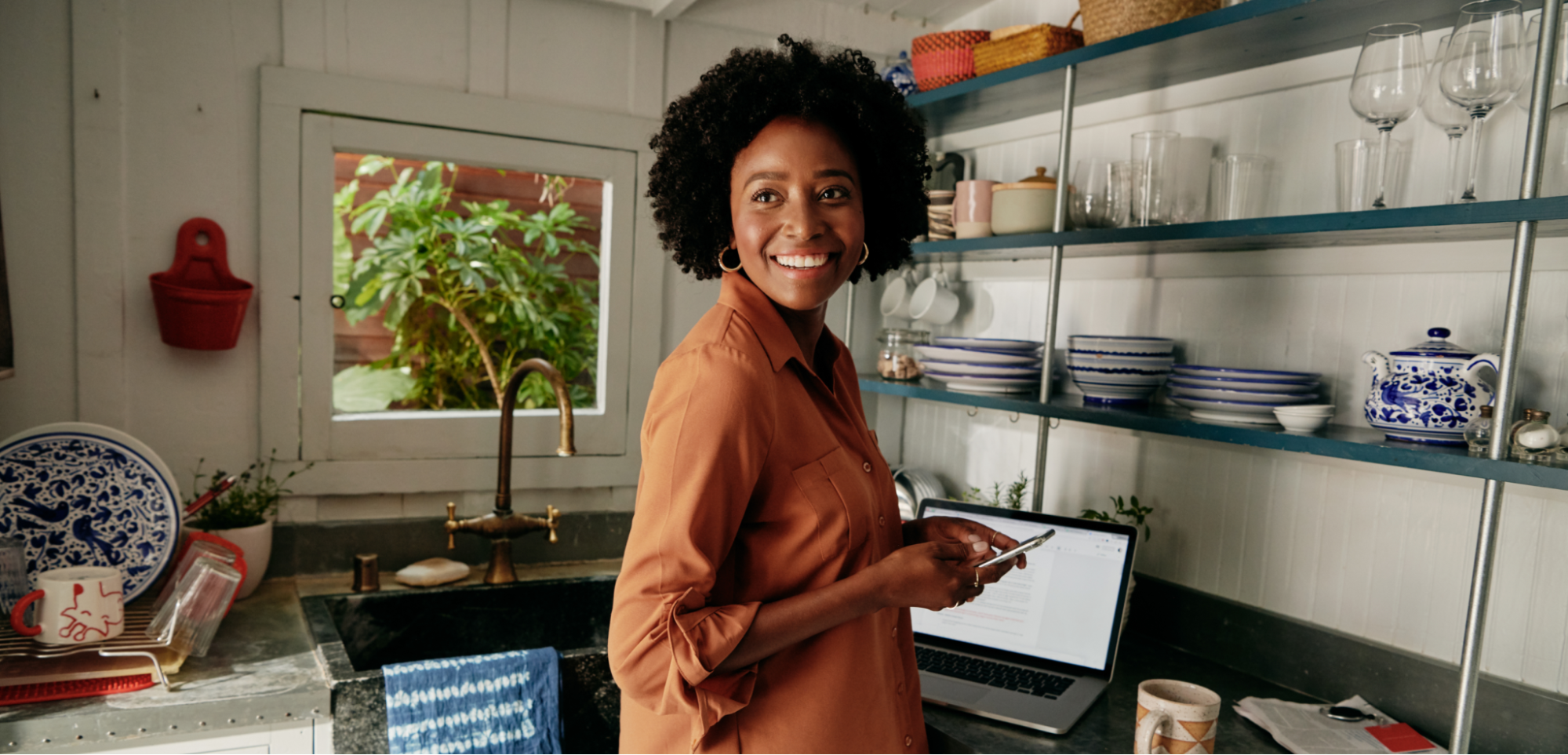 Une personne souriante, un téléphone à la main, se tient devant un ordinateur portable ouvert sur le comptoir d'une cuisine.