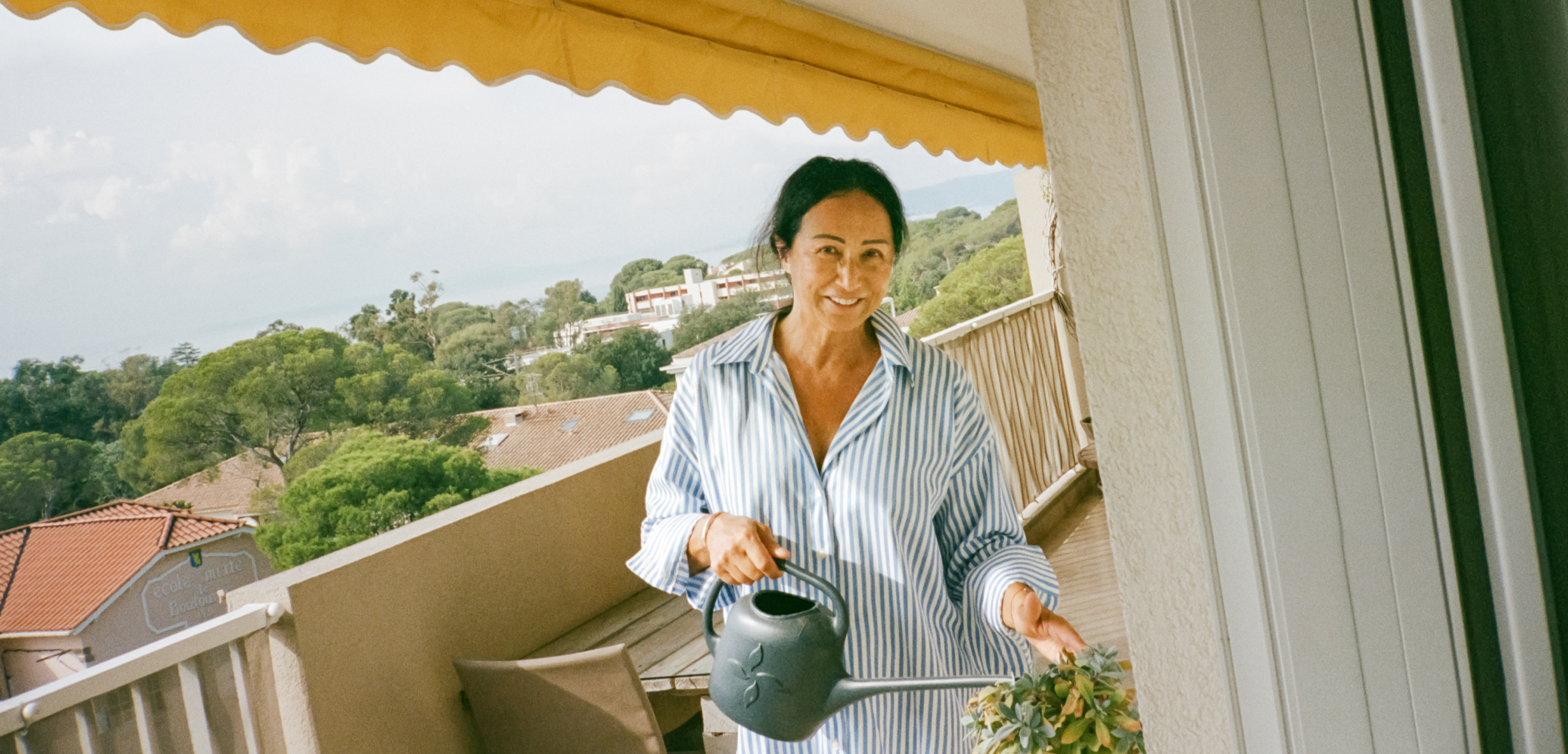 A person holding a watering can on a deck smiles, with trees and sky in the background.