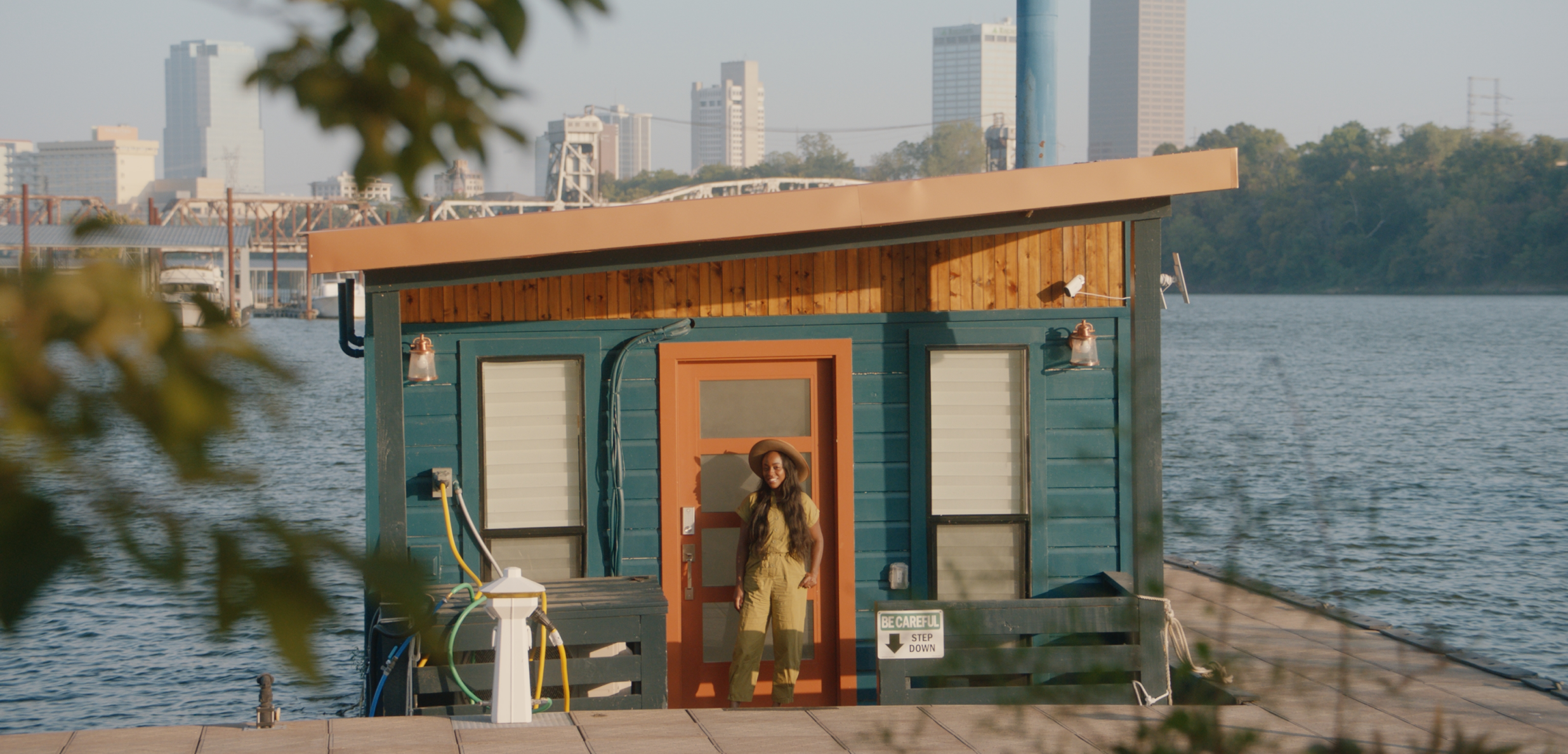 Un anfitrión sonríe frente a una casa flotante con vista al agua, los árboles y el paisaje urbano a lo lejos.