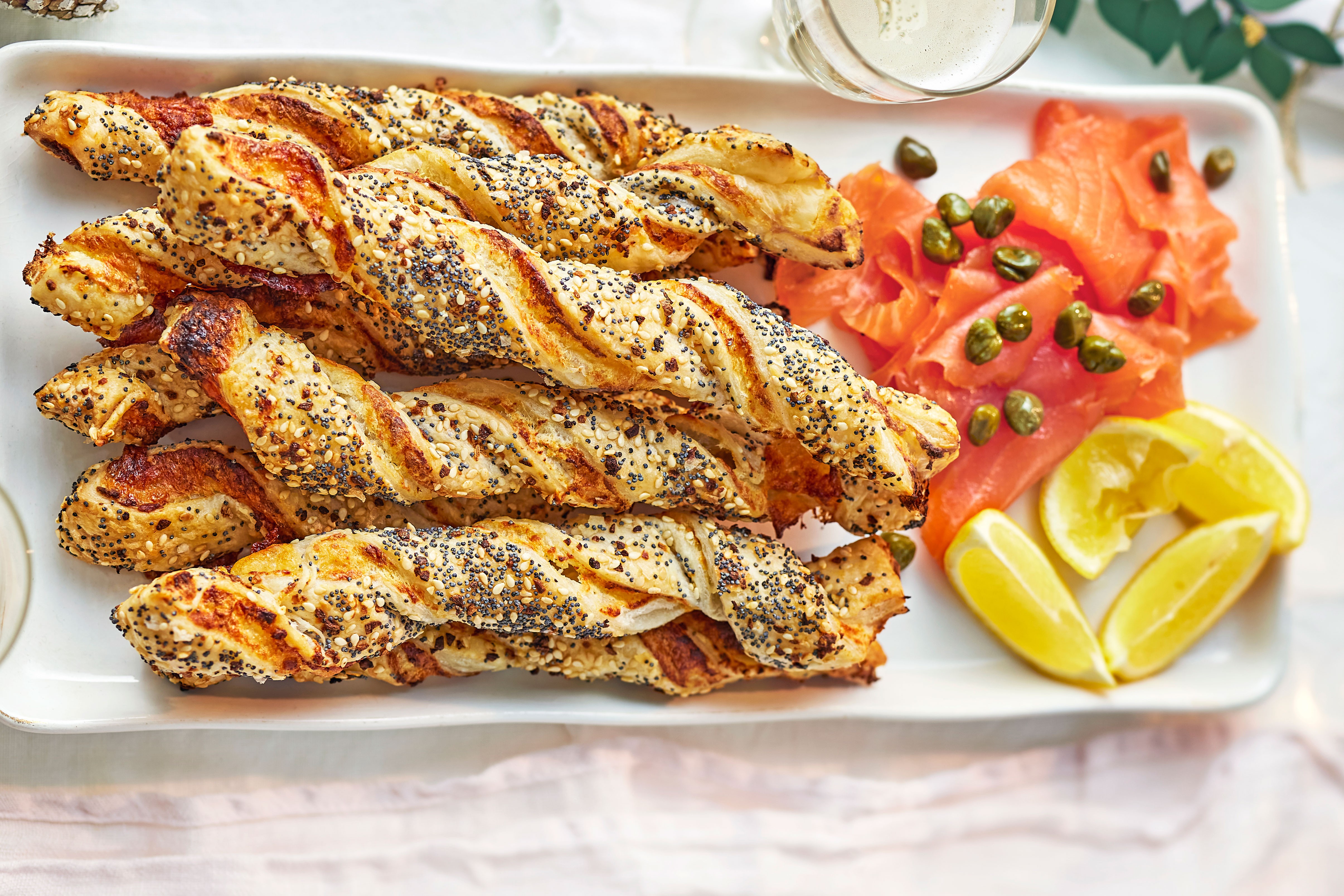 Cheese straws topped with seeds next to smoked salmon, capers and lemon wedges