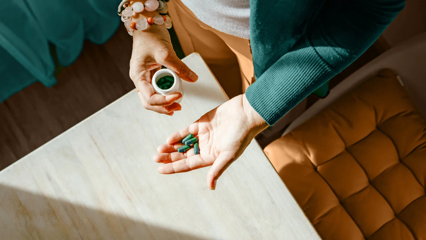 Mature woman standing at a table with green pills in her hand.