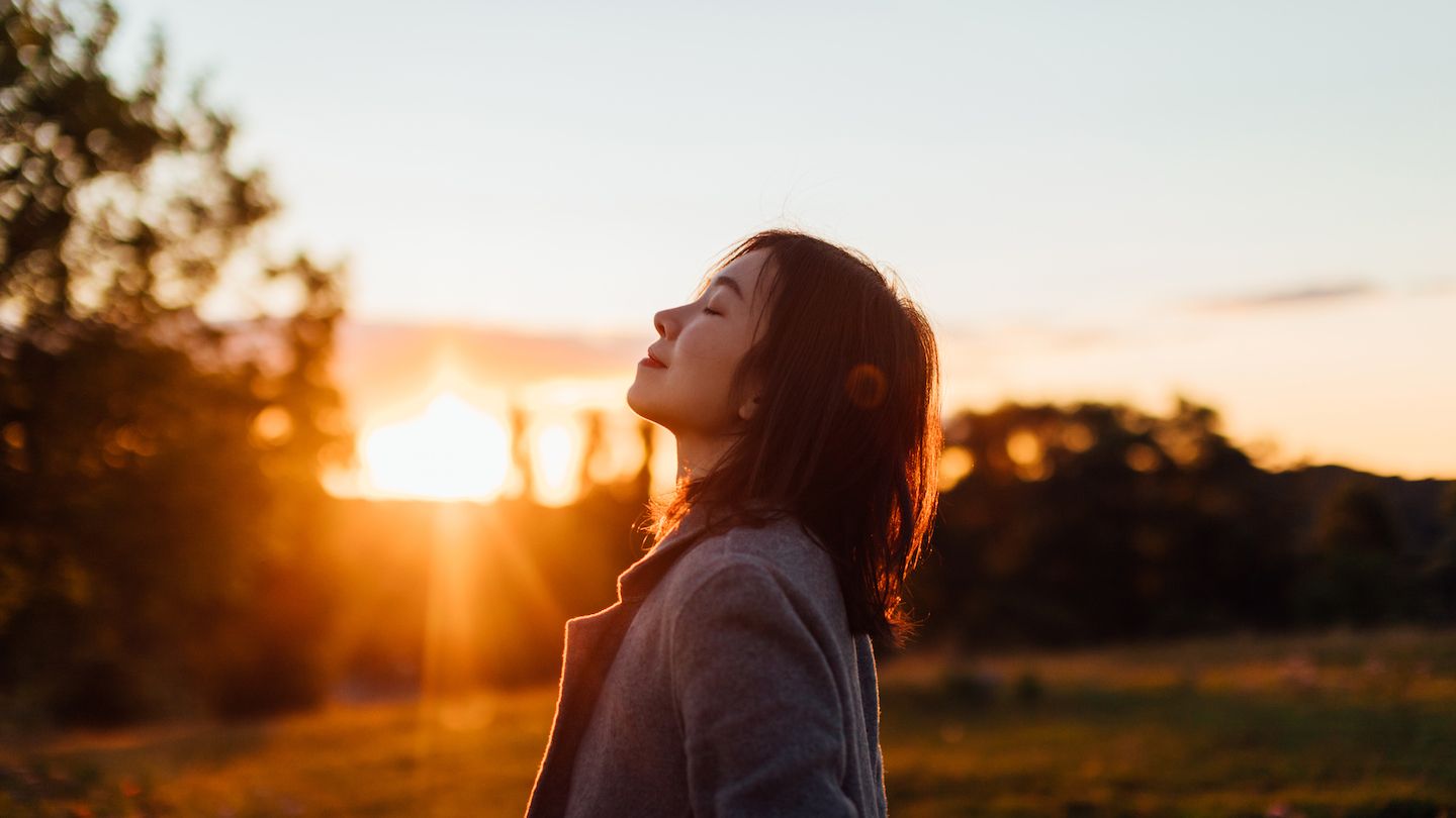 a young woman with brown hair faces a sunset with her eyes closed