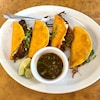 Quatre tacos dans une assiette avec des quartiers de lime et un petit bol de bouillon.