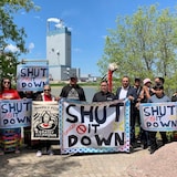 Protesters stand in front of the Dryden Paper Mill, holding signs saying "Shut it down" and "Justice for Grassy Narrows".