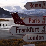 A signpost in the foreground points towards various destinations at the Kangerlussuaq Airport in Greenland as passengers walk to an airplane in the background.  