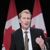 The minister speaks at a press conference in front of Canadian flags.