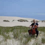 Une violoncelliste joue de son instrument dans un champs avec une dune de sable et des herbes.