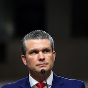 Pete Hegseth, habillé d'un veston, fait face, impassible, à un micro.