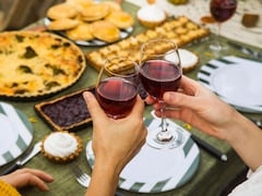 Deux personnes, dont on ne voit que les mains, trinquent avec leur verre de vin rouge. À l'arrière-plan, une table est remplie de nourriture. 