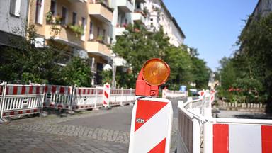 Rot-weisse Baustellenbegrenzungen sperren die Parkplätze entlang einer Straße ab, im Vordergrund steht eine orangene Warnlampe.