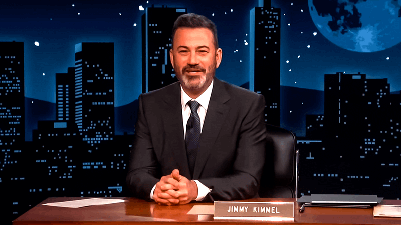 Jimmy Kimmel sitting at the Jimmy Kimmel Live! desk in a suit with a city backdrop