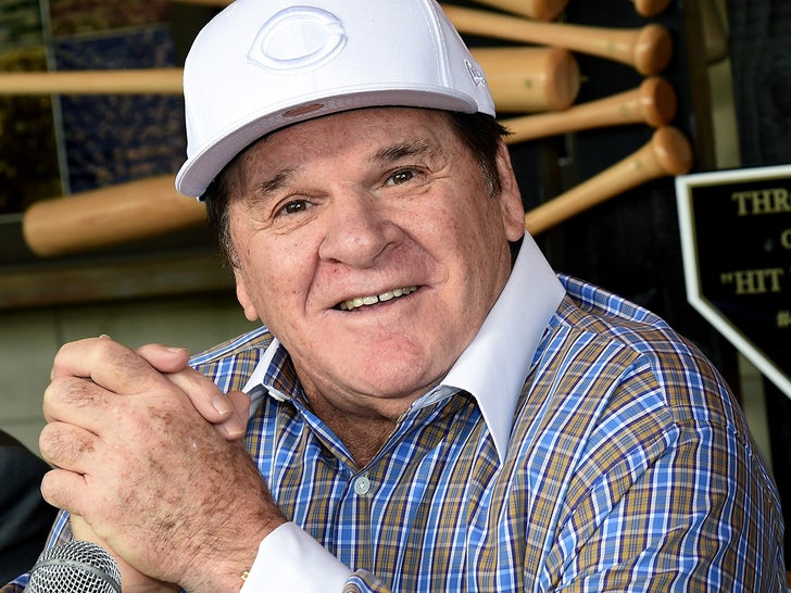 Pete Rose smiling while wearing a shirt with contrast white cuffs and collar, and a white Cincinnati Reds hat.