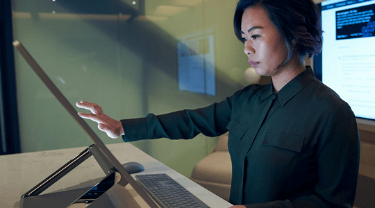 Πλαϊνό προφίλ μιας γυναίκας που φοράει σκούρο μπλουζάκι σε ένα αχνό γραφείο με κύλιση ή εργασία σε Surface Studio της Microsoft.