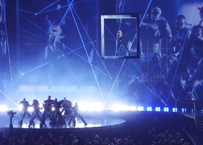 Dancers perform onstage while a woman sings in a suspended box