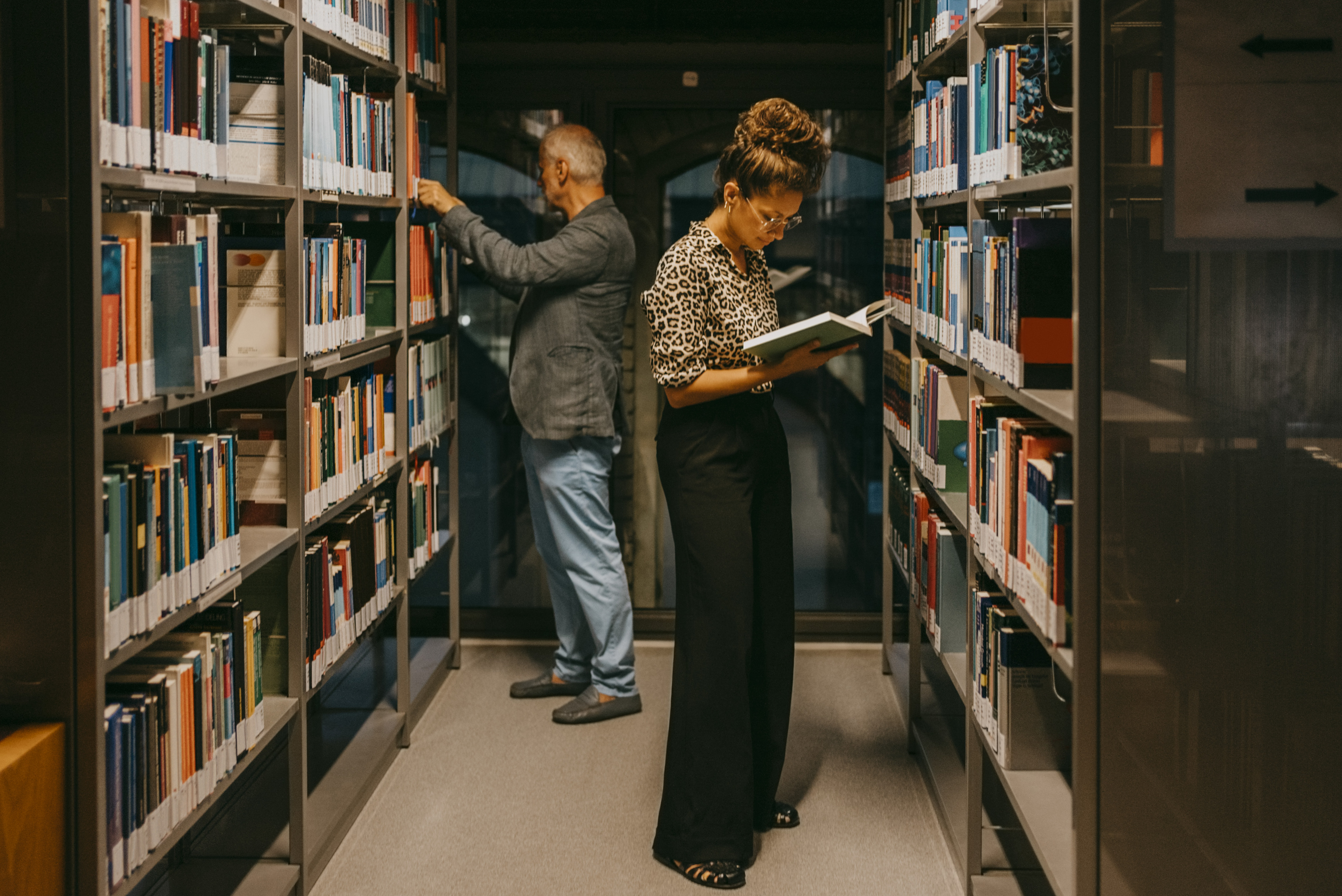 Two people in a library aisle, one reading a book and the other browsing shelves