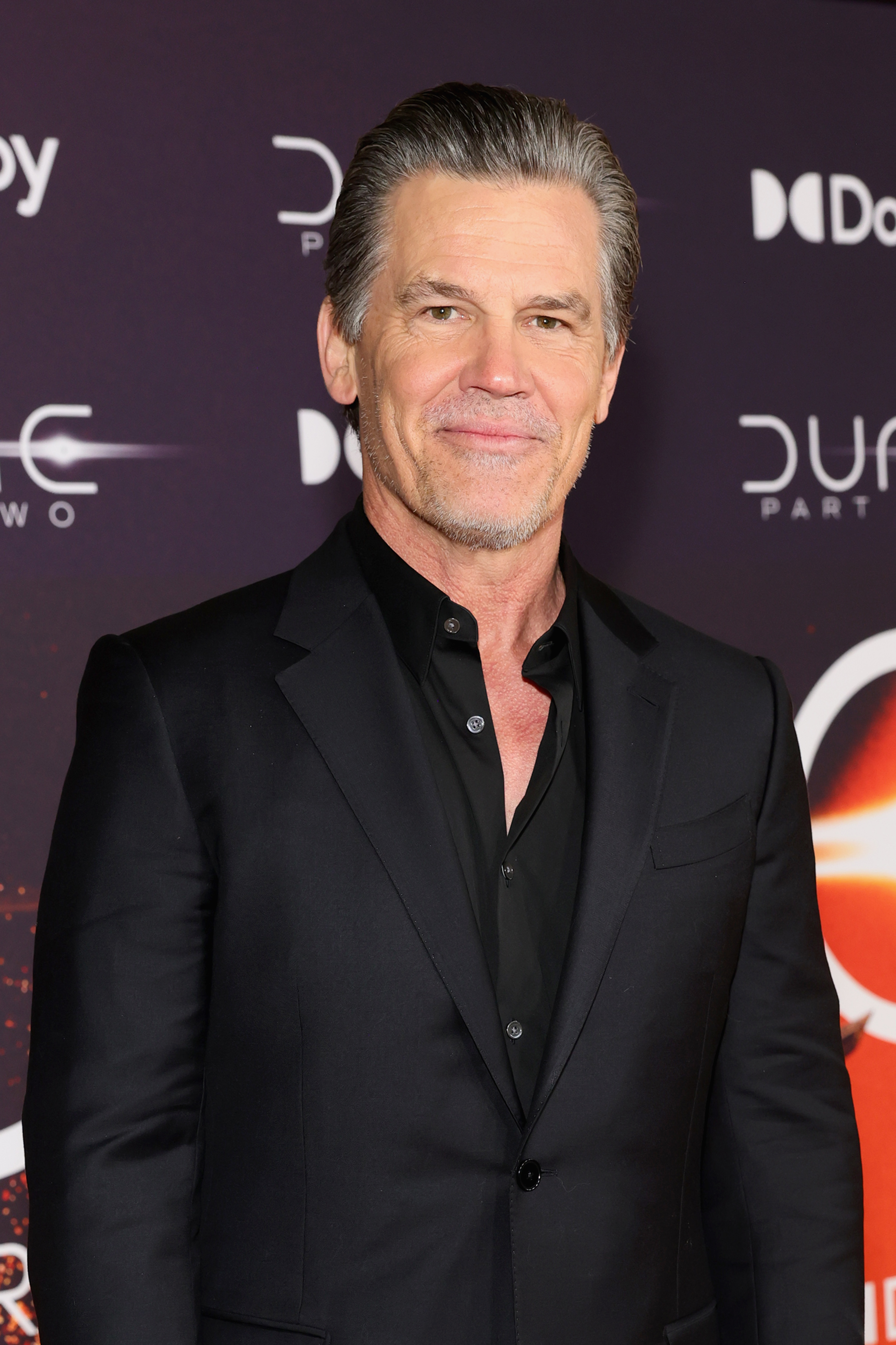 Josh Brolin on a red carpet, wearing a black suit with a black dress shirt, attending an event for &quot;Dune: Part Two.&quot;