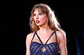 Taylor Swift performs on stage wearing a sparkling, fringed bodysuit with matching boots and fishnet stockings. The background features stage lights