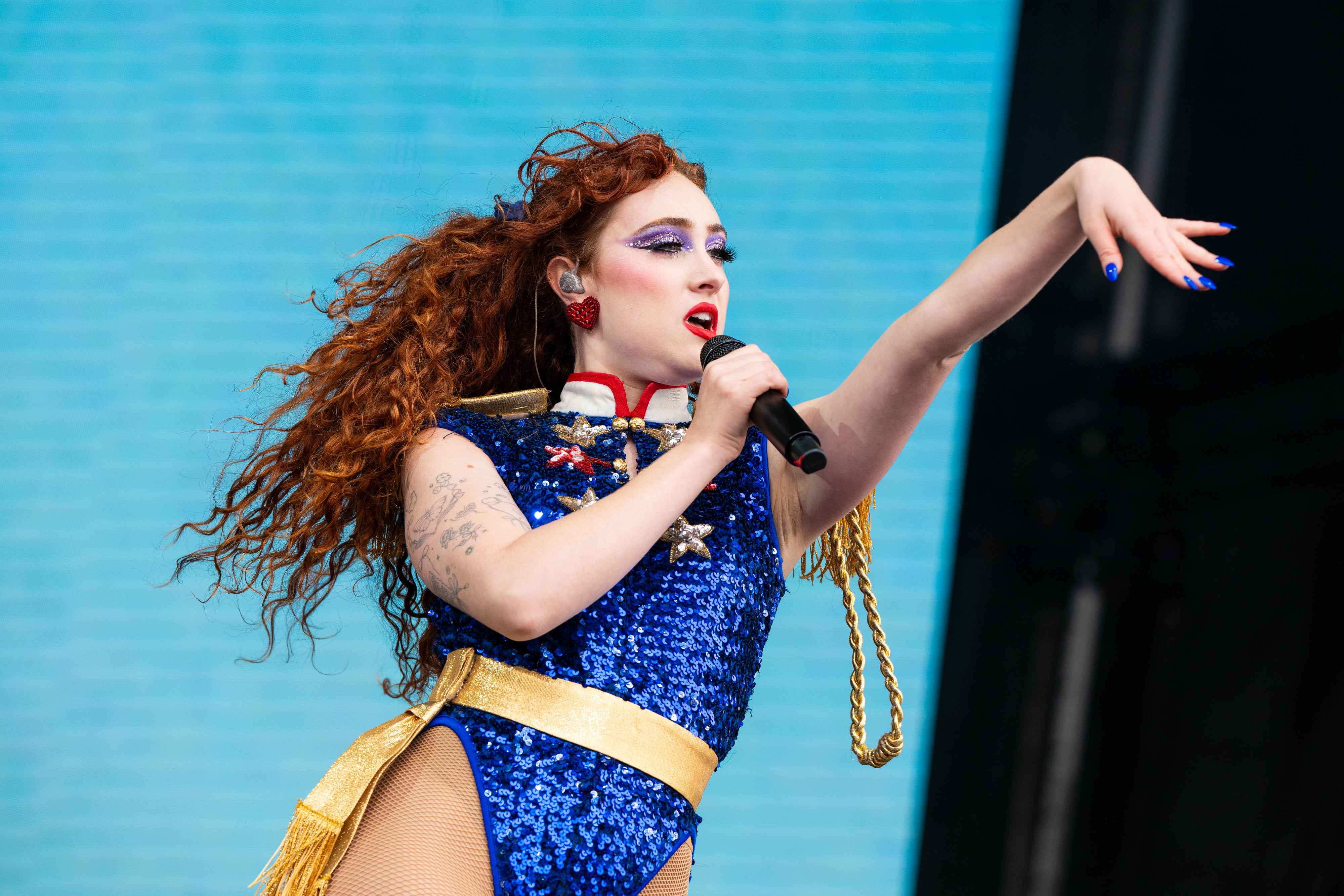 Chappell Roan with long curly hair on stage, wearing a sparkling bodysuit with accents, holding a microphone mid-performance