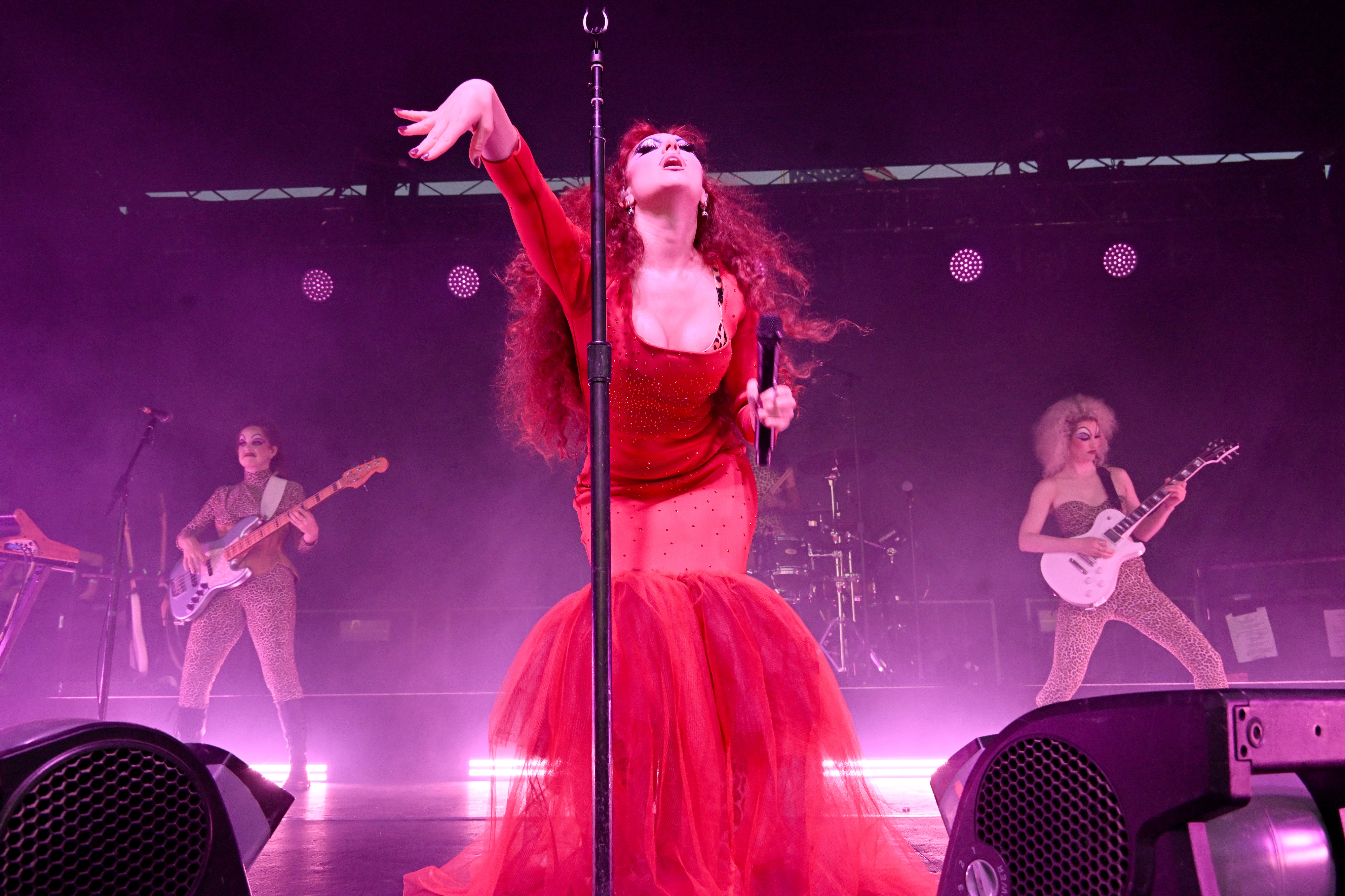 Chappell Roan in a dramatic outfit with a mermaid-style skirt sings energetically on stage. Backup musicians with instruments are in the background