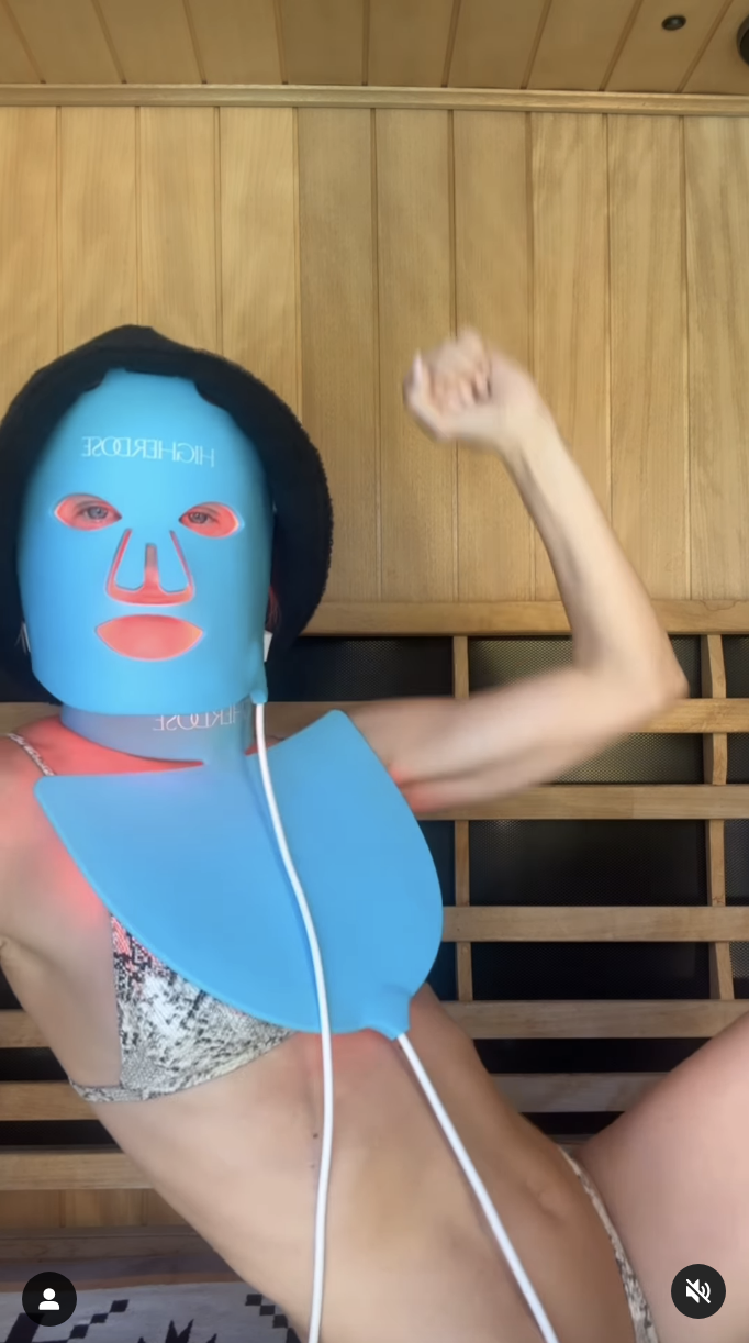 Julianne wearing a LED face mask and matching neck piece, posing energetically in a bikini, inside a wooden sauna-like room