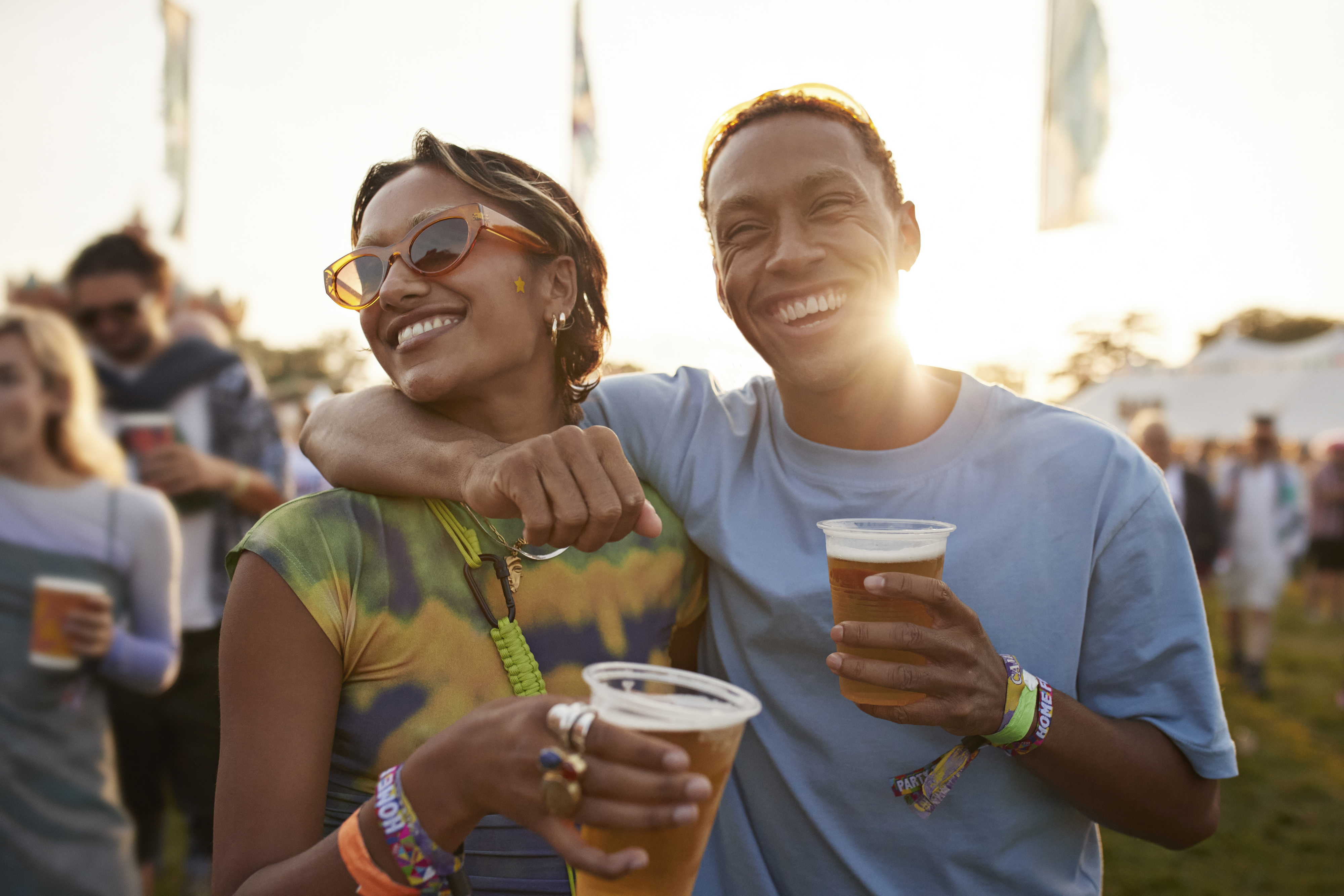 A happy couple at an outdoor event, each holding a cup, with the sun setting behind them, creating a warm and joyful atmosphere