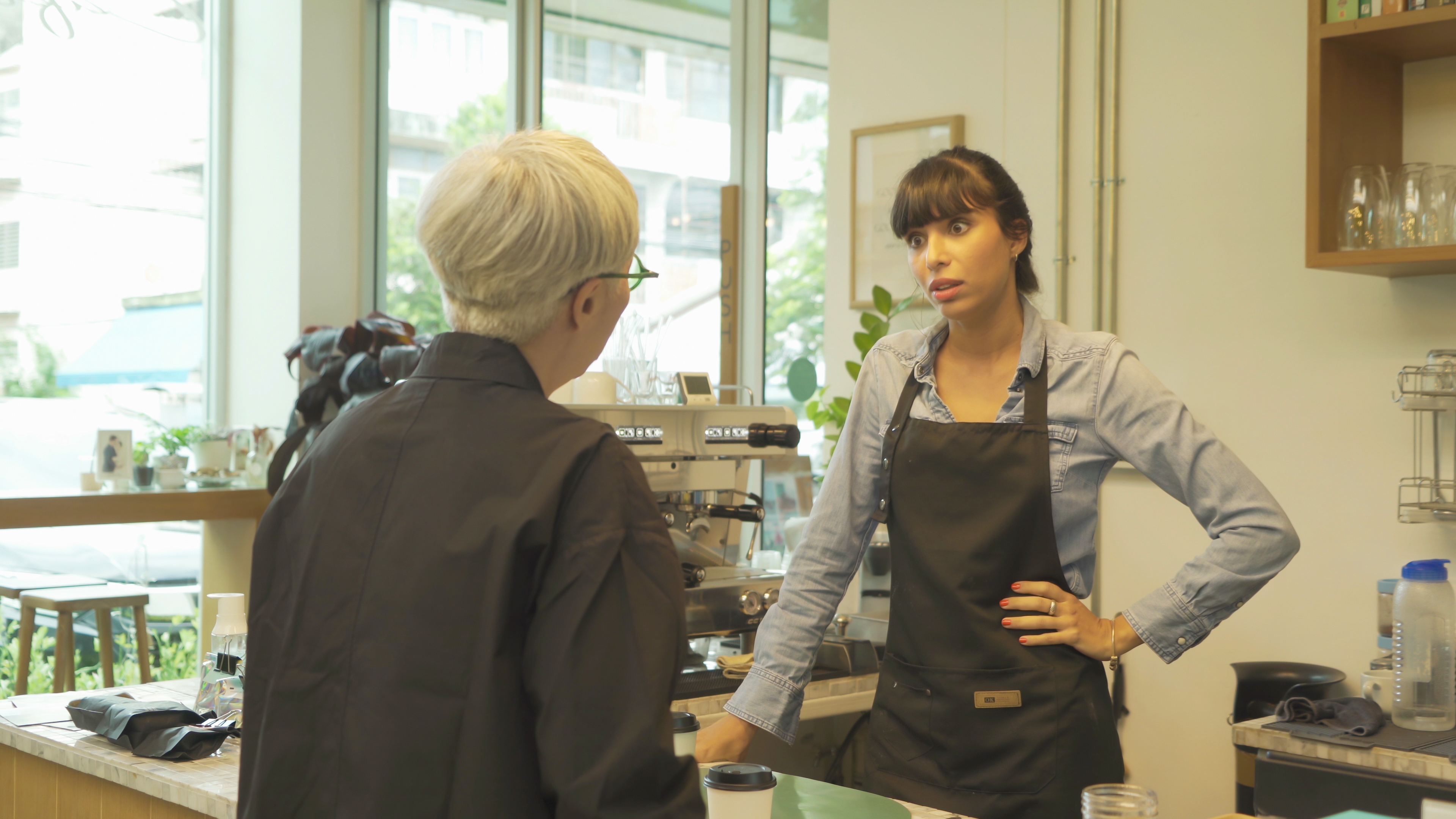 Two people in a coffee shop, one with gray hair facing a counter, the other with dark hair and apron, standing behind the counter, talking