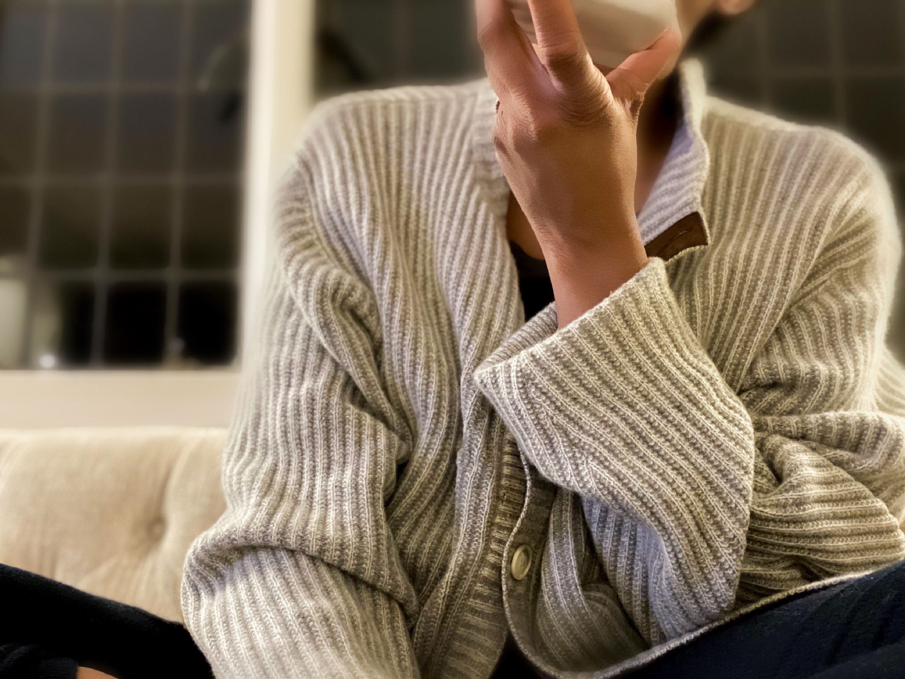 Person in a textured cardigan, seated and holding a cup near their face. Room is softly lit; window in the background