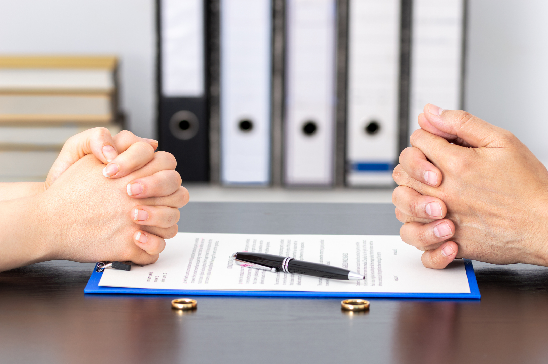 Two hands clasped together face each other over divorce papers, a pen, and two rings