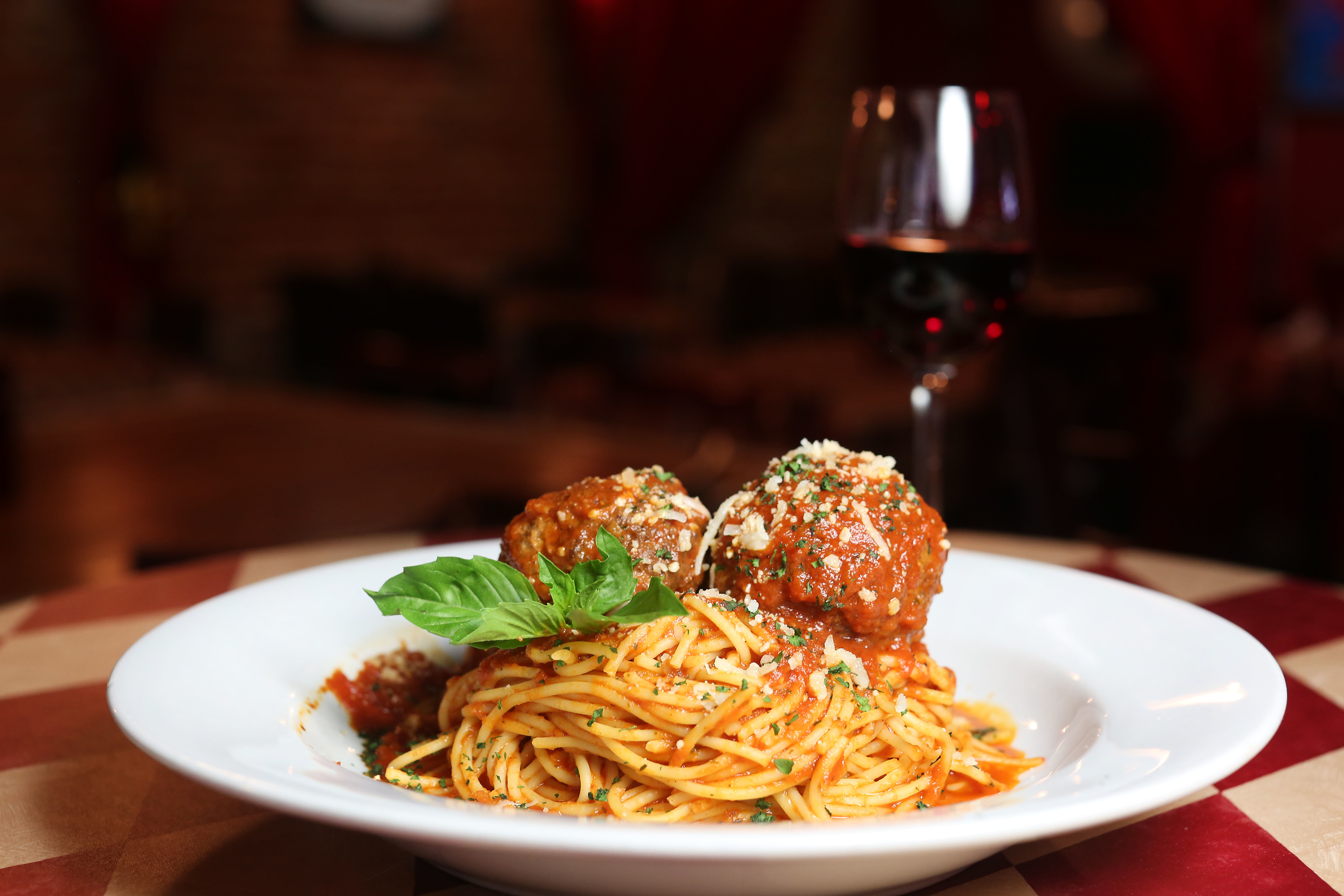 Plate of spaghetti with meatballs and herbs, garnished with cheese, next to a glass of red wine on a checkered tablecloth