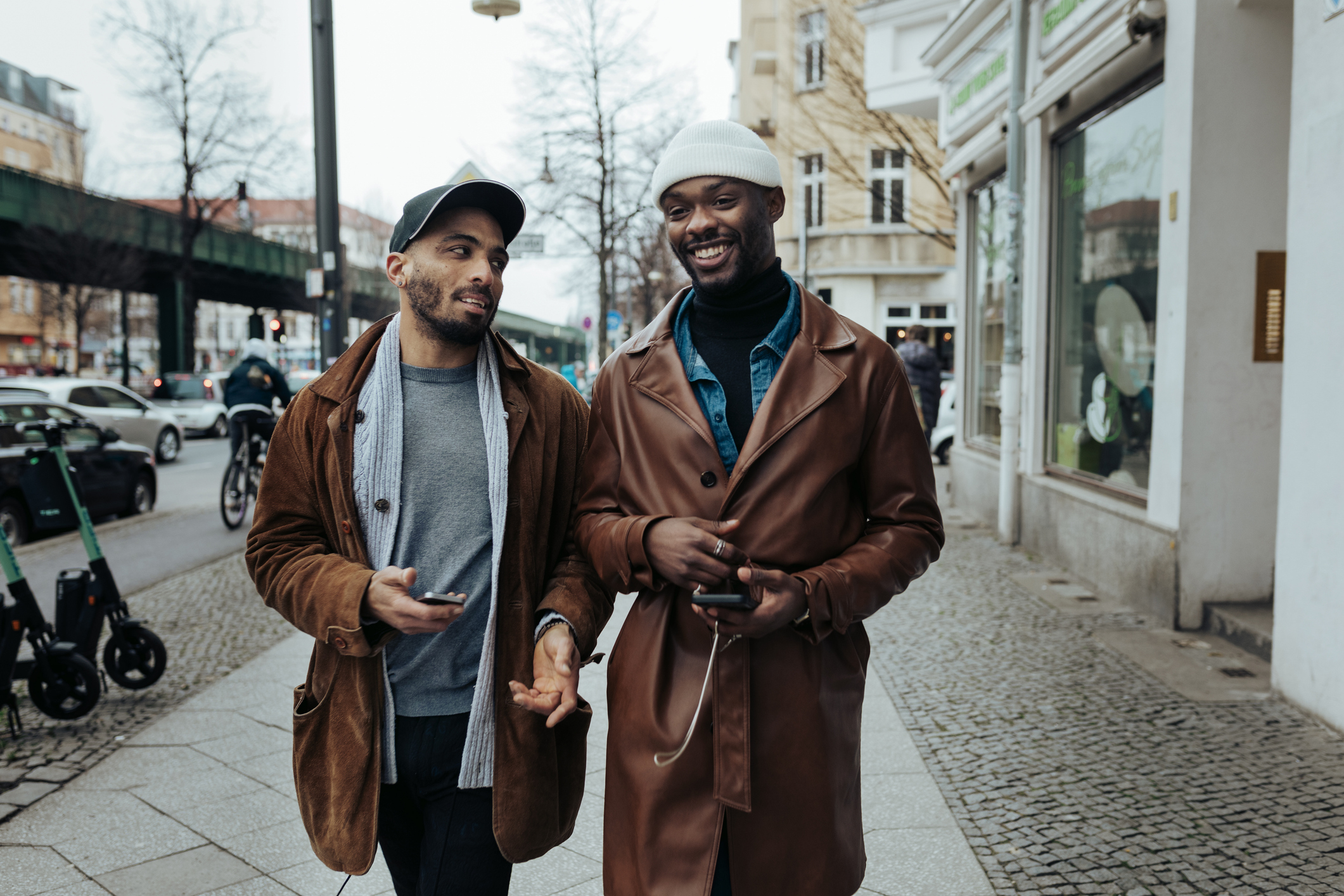 Two people walking and smiling on a city sidewalk, both wearing stylish coats and hats, holding phones and chatting