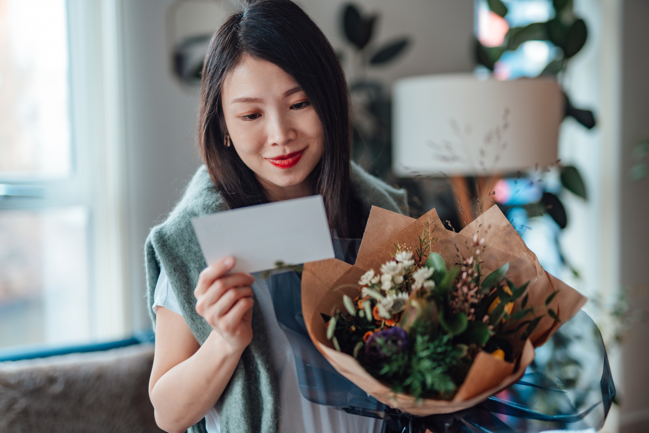 Woman smiling at a greeting card, holding a bouquet of flowers, in a cozy home setting