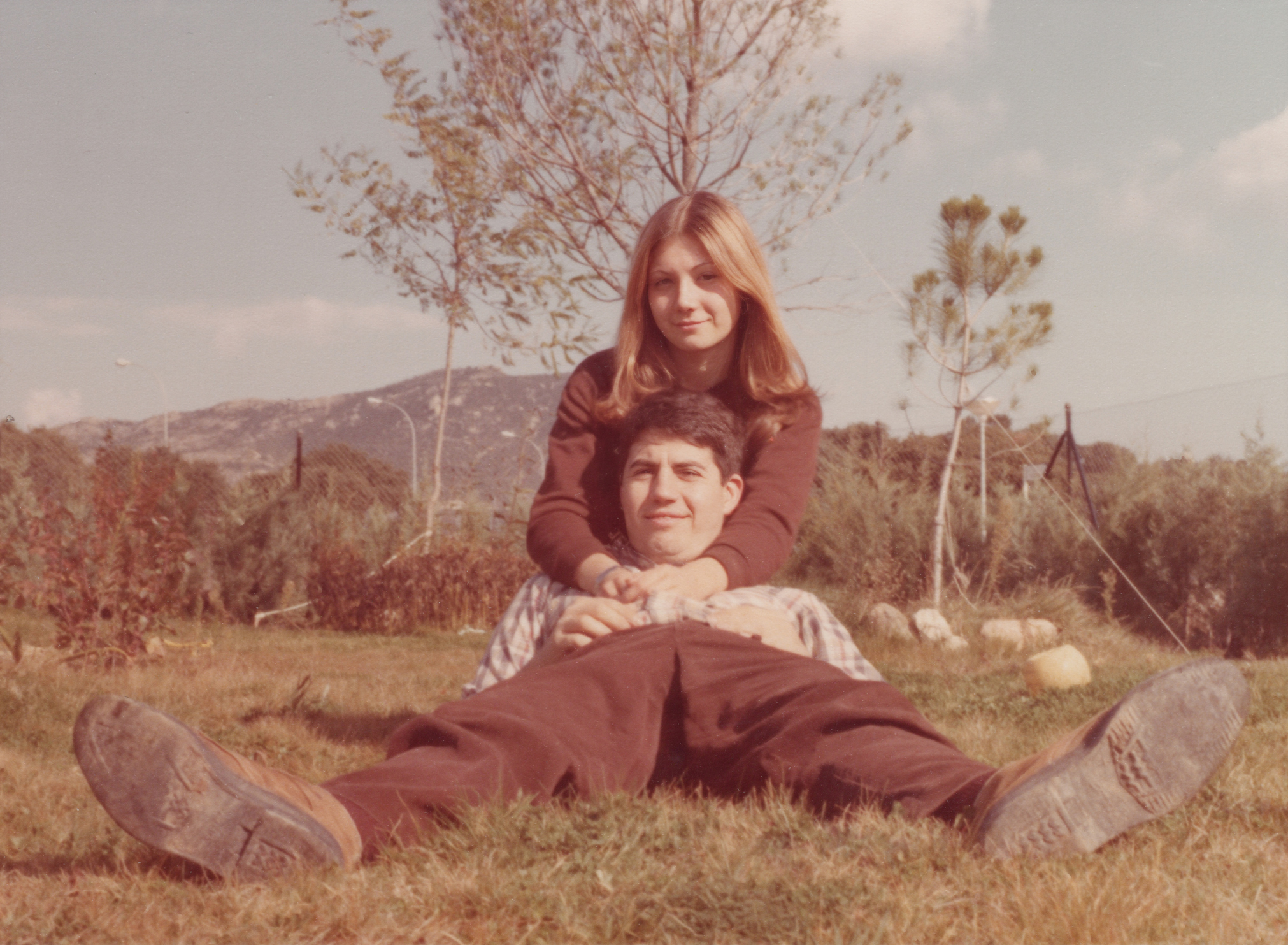 A couple sits closely on grass outdoors, with the woman embracing the man from behind. They both appear relaxed and happy