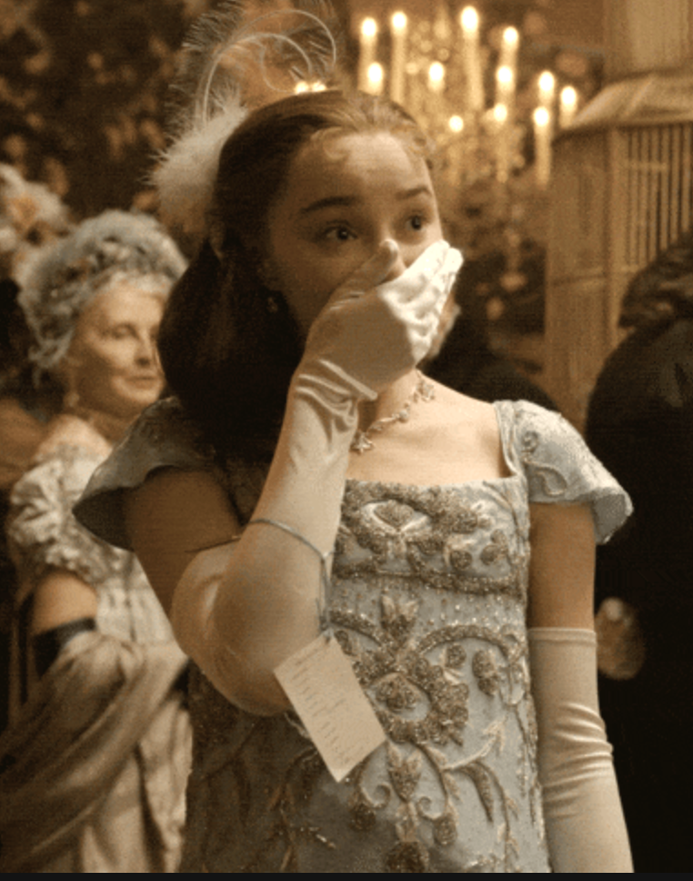 Person in an elegant, embroidered gown with gloves and a feathered headpiece, covering their mouth in surprise at a formal event