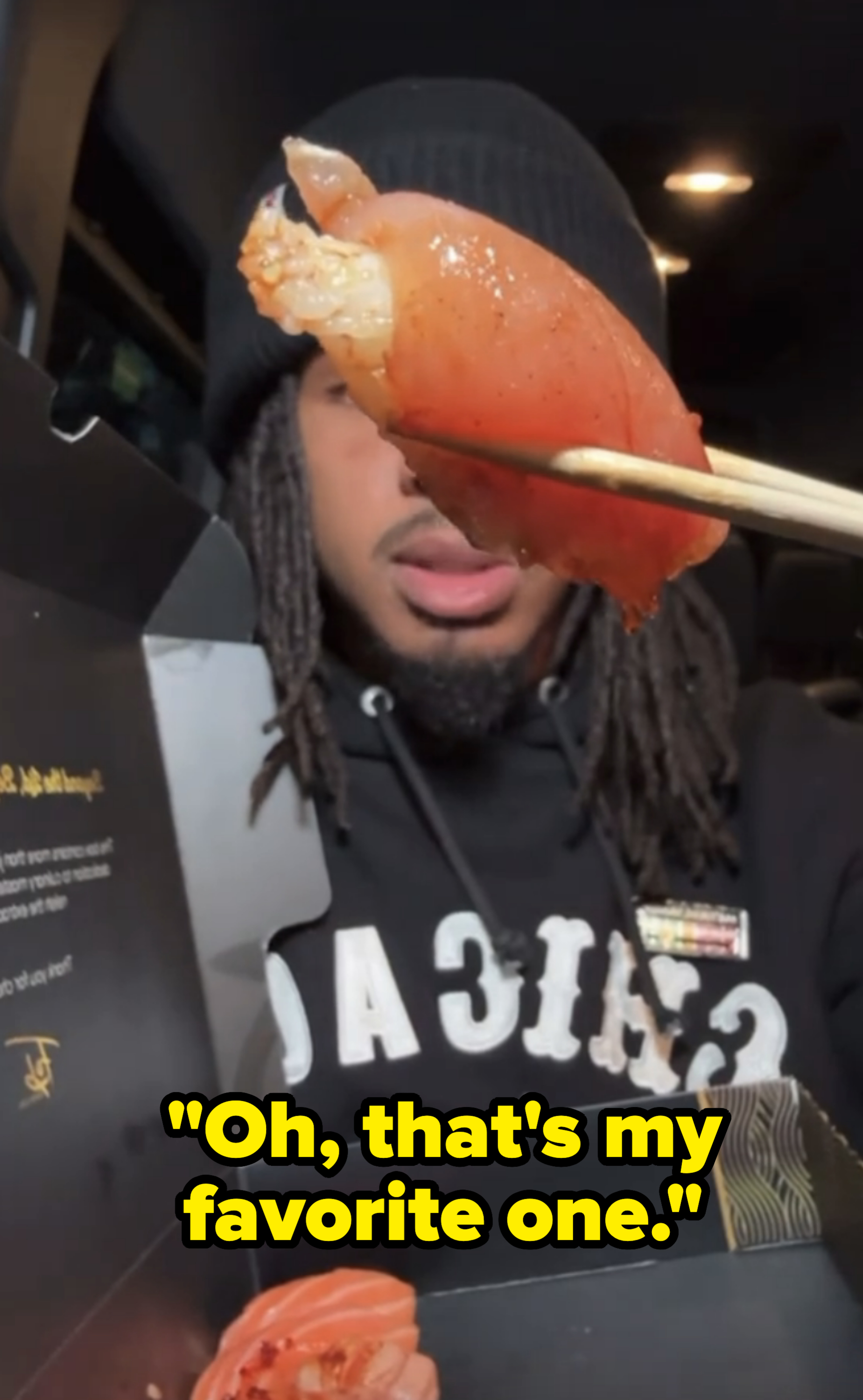 Person holding sushi with chopsticks, ready to eat in a dimly lit setting. They wear a beanie and a dark hoodie