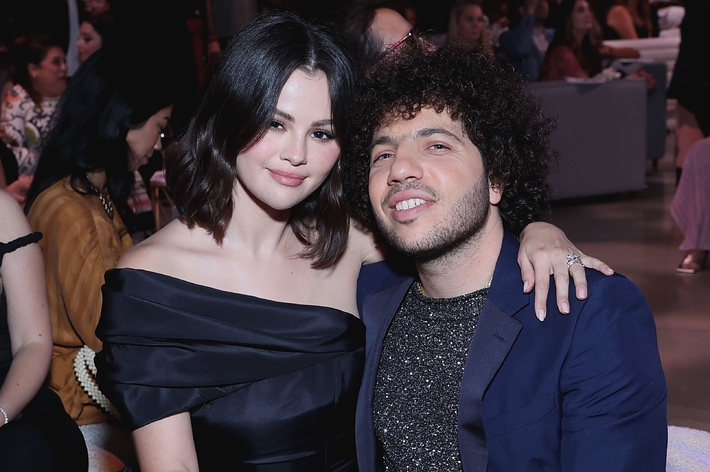 Two people sitting; one holds a number 5 paddle. The person on the left wears a stylish off-shoulder dress. They are at an event with seated guests