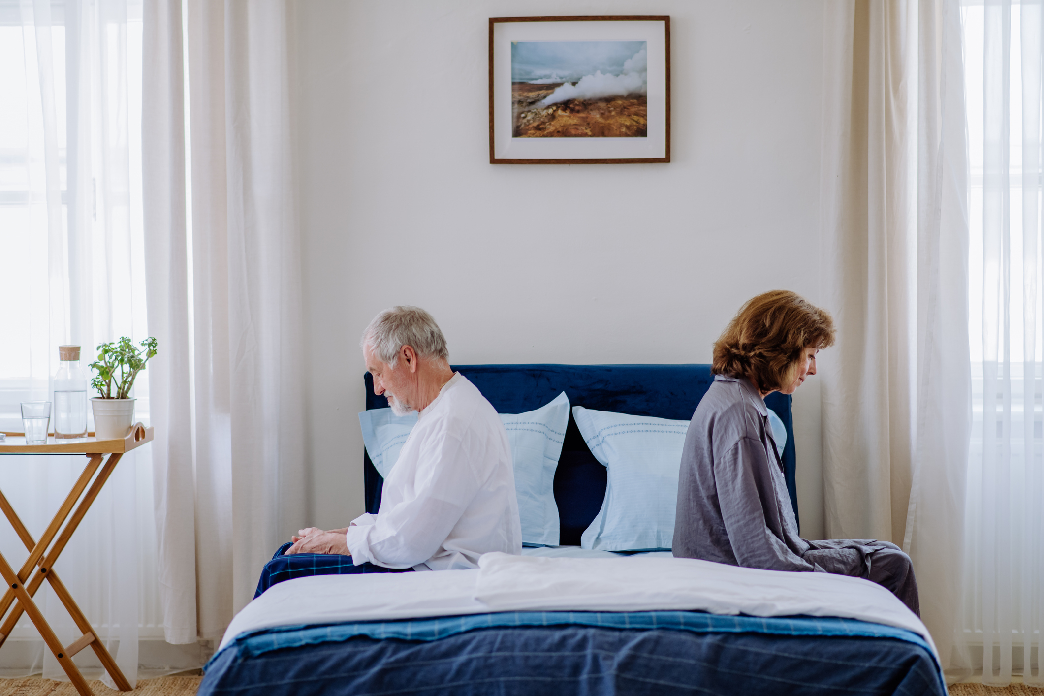 A couple sits back-to-back on a bed, appearing distant and thoughtful, conveying themes of relationship tension or contemplation