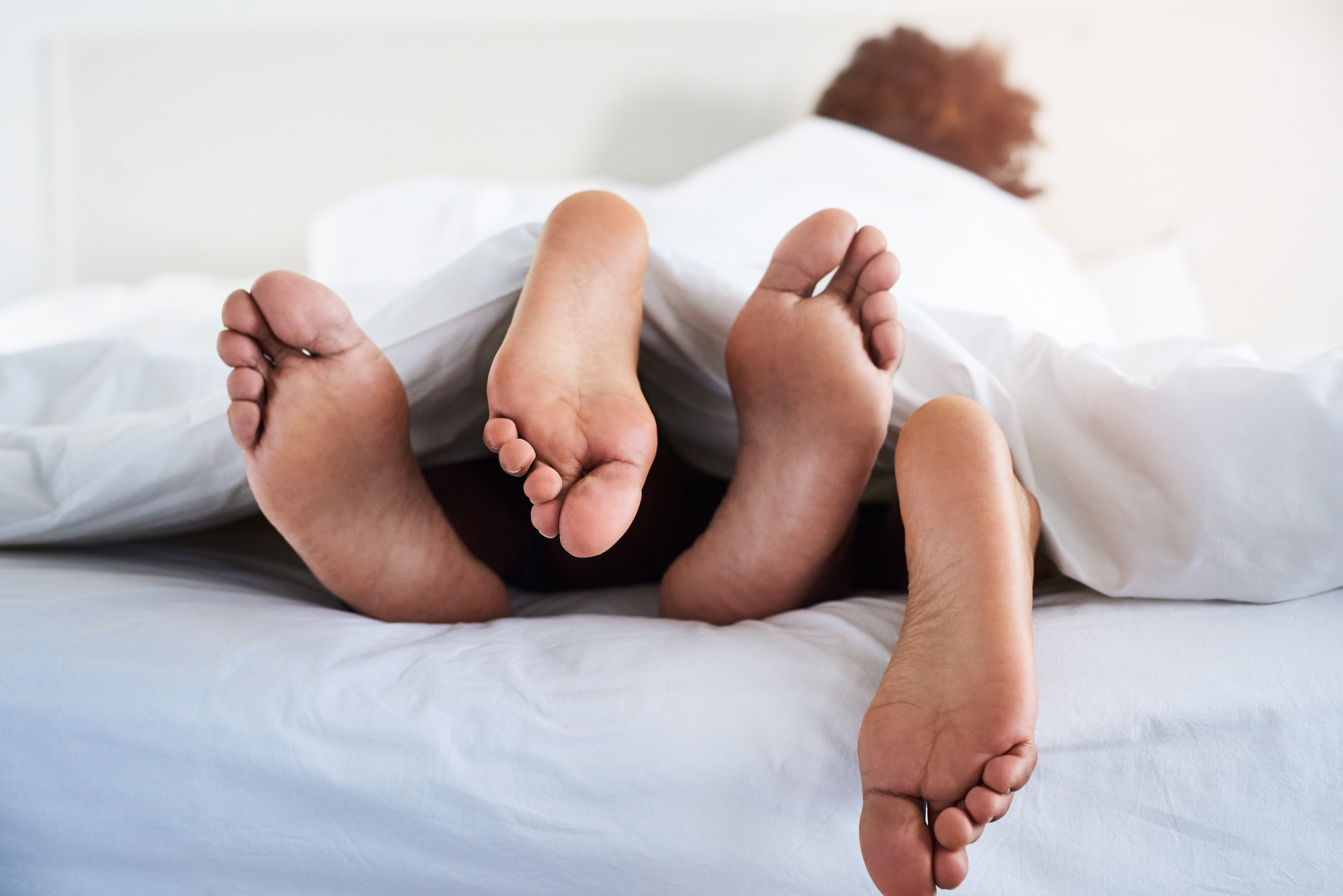 Two pairs of feet stick out from under white bed covers, suggesting intimacy and romance