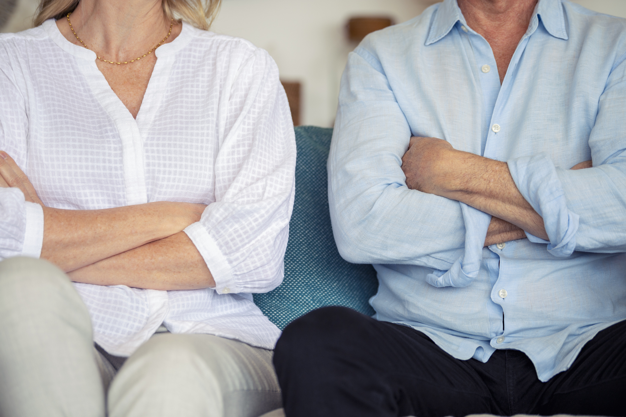 A couple sits with their arms crossed, wearing relaxed, casual clothing. The image conveys tension or disagreement