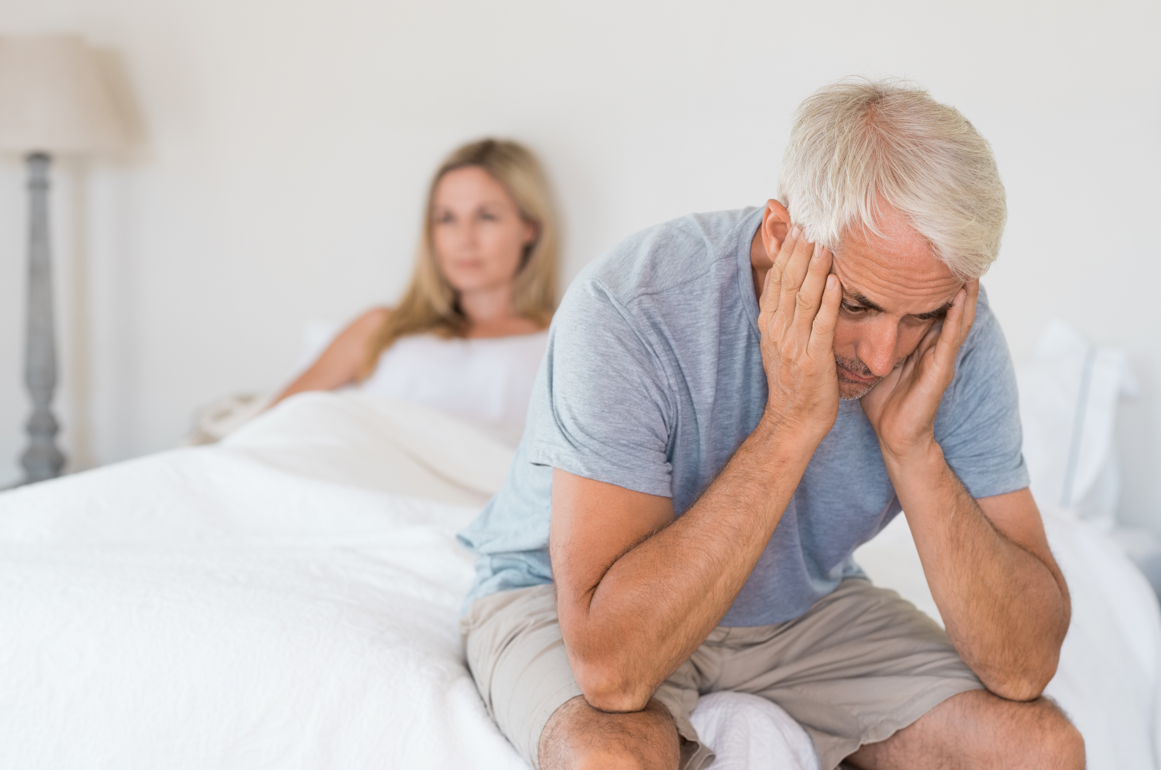 A man sits on a bed looking distressed, while a woman sits in the background looking thoughtful