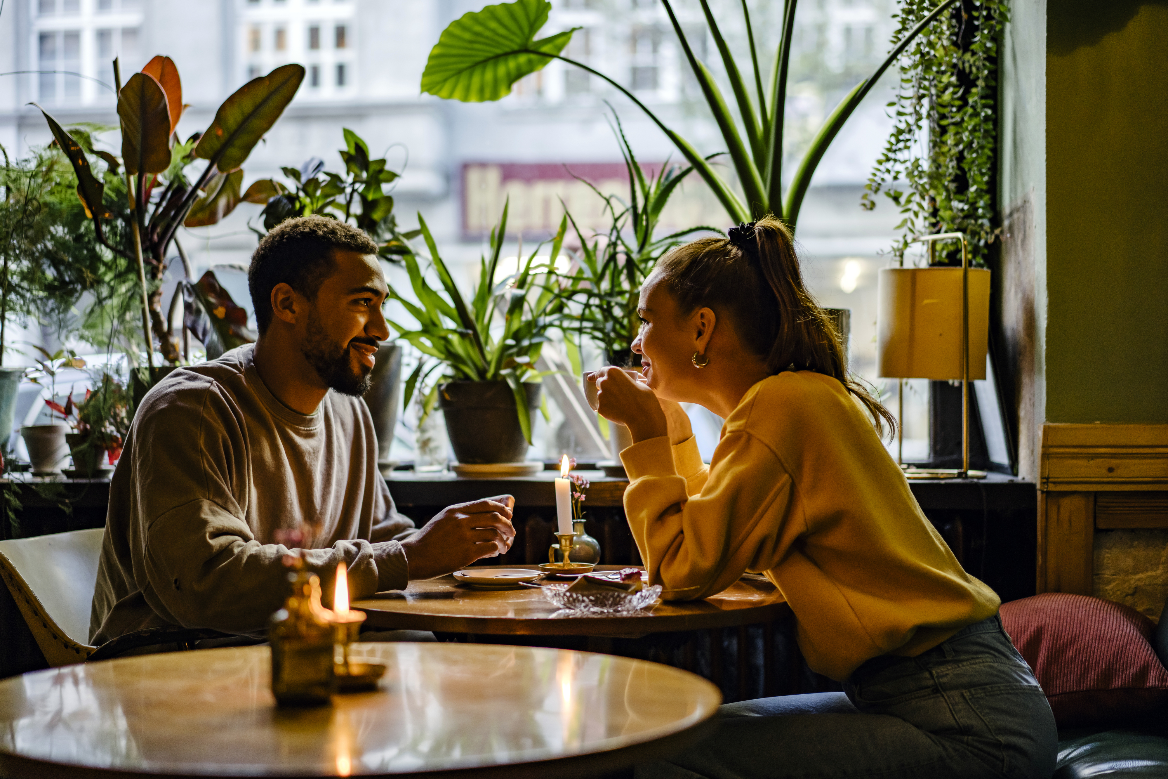 A couple enjoys a romantic dinner at a cozy restaurant, surrounded by plants and candlelight, deep in conversation and smiles