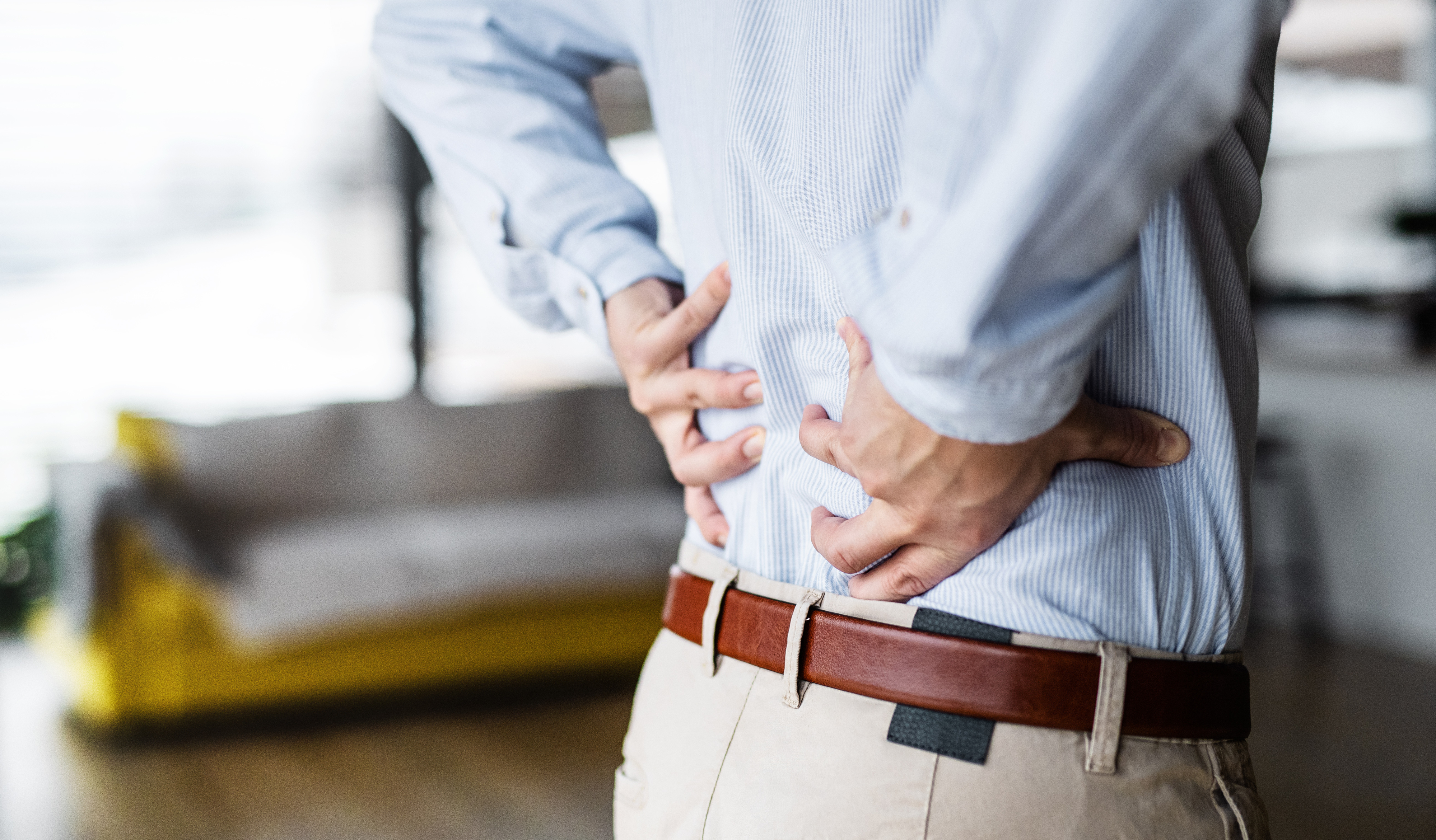 Person in a button-up shirt holding their lower back, indicating discomfort, in a home setting