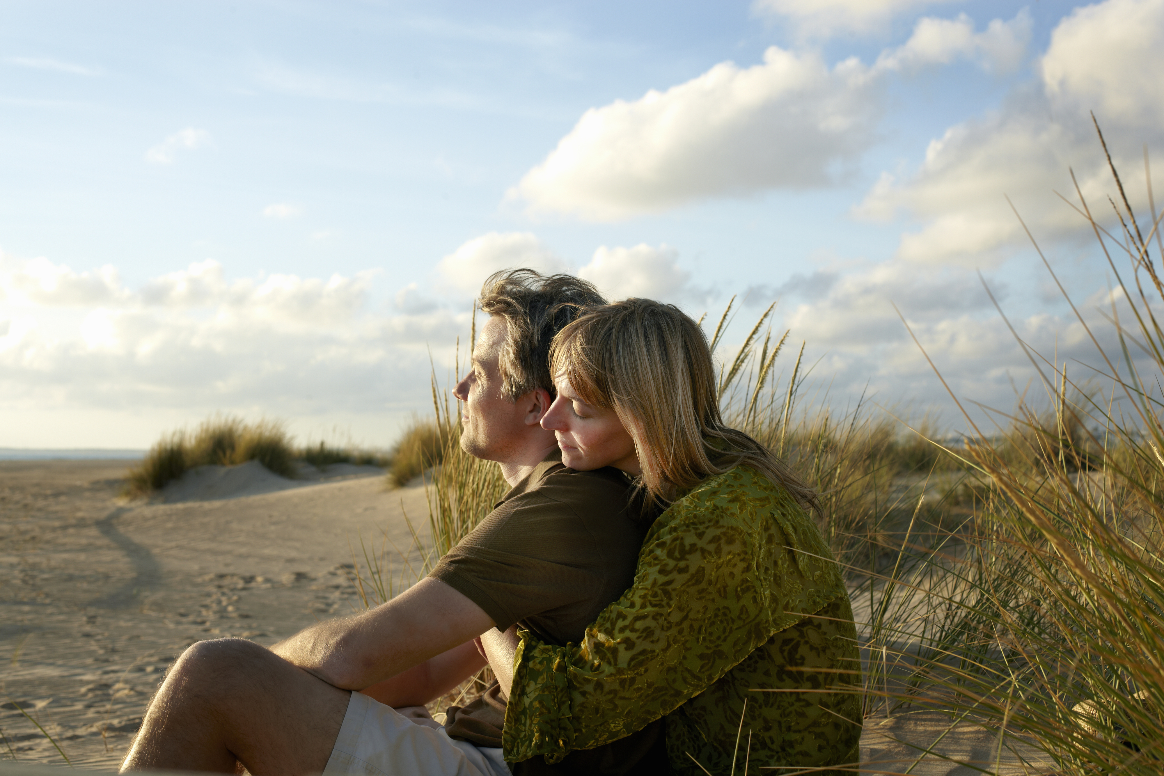 A couple sits closely on a beach surrounded by dunes, gazing thoughtfully at the horizon