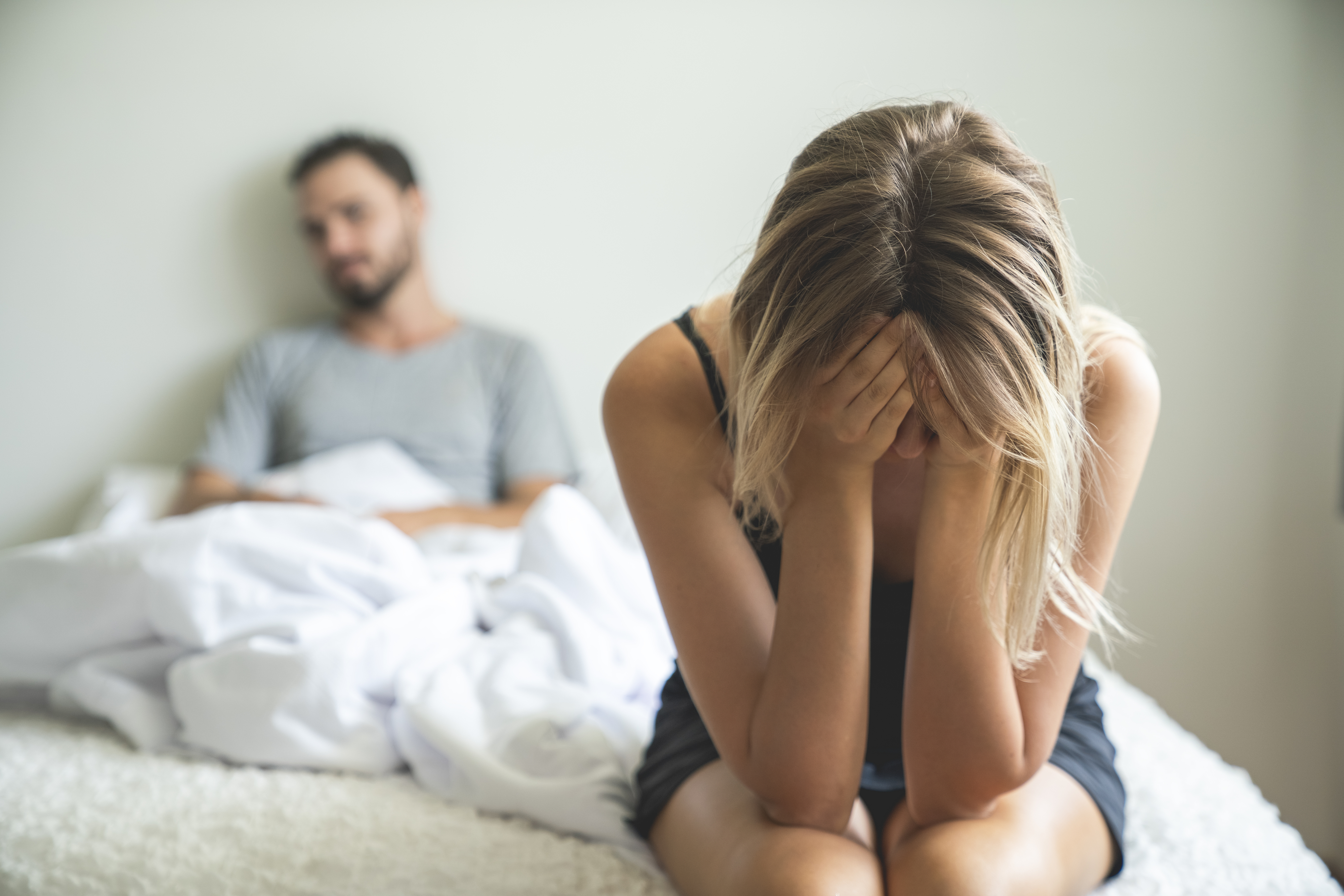 A woman sits on a bed with her head in her hands, appearing upset, while a man sits in the background looking away